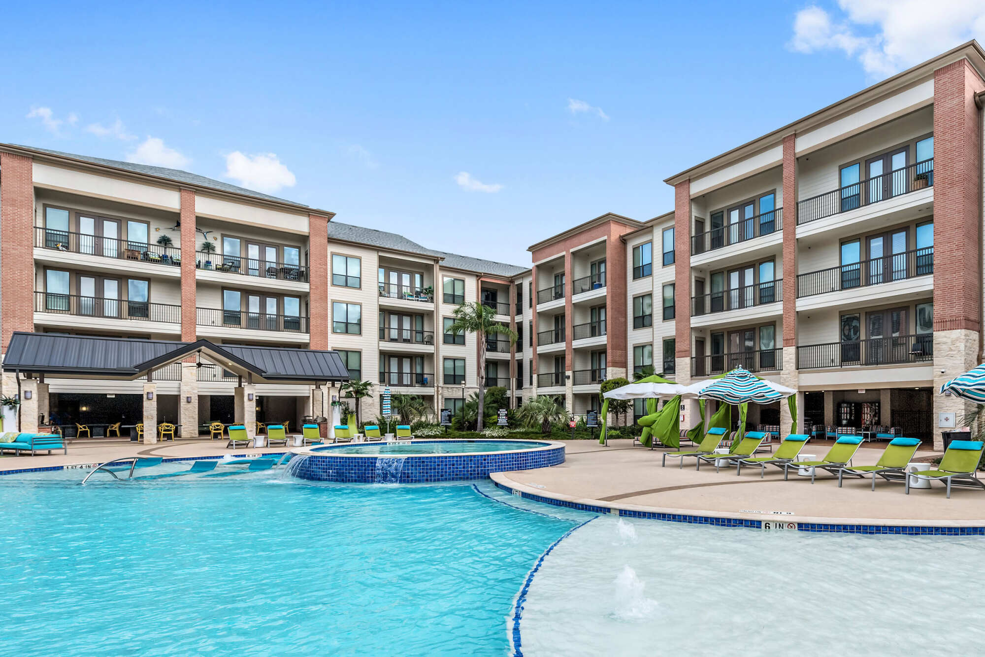 Resort-style courtyard pool with a tanning shelf and water bubblers, lounge chairs, teal umbrellas, cabanas, and brick-and-stucco apartment buildings under a blue sky.