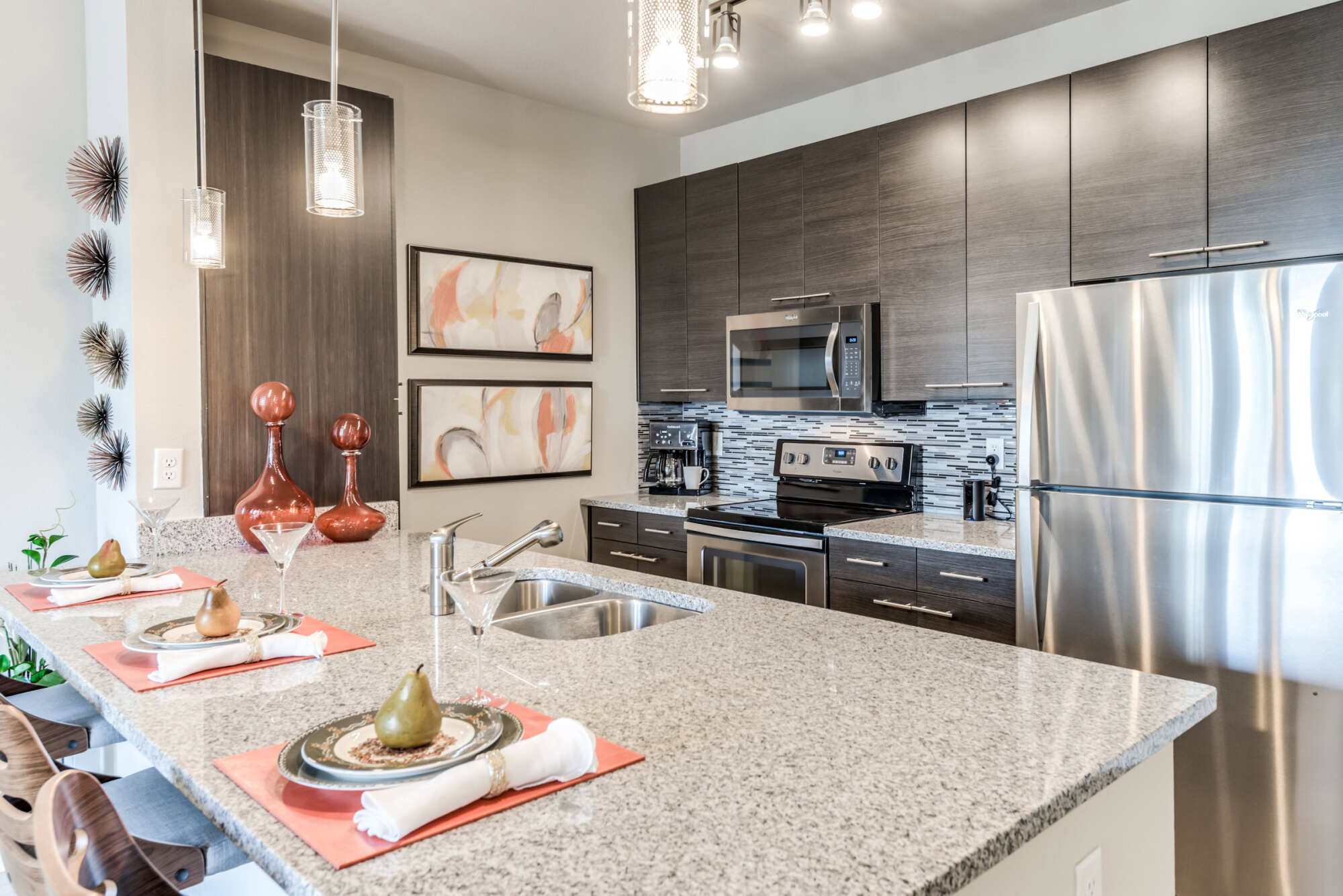 Apartment kitchen featuring a granite island, dark wood cabinets, glass-tile backsplash, and stainless steel appliances, with a staged place setting and pendant lighting.