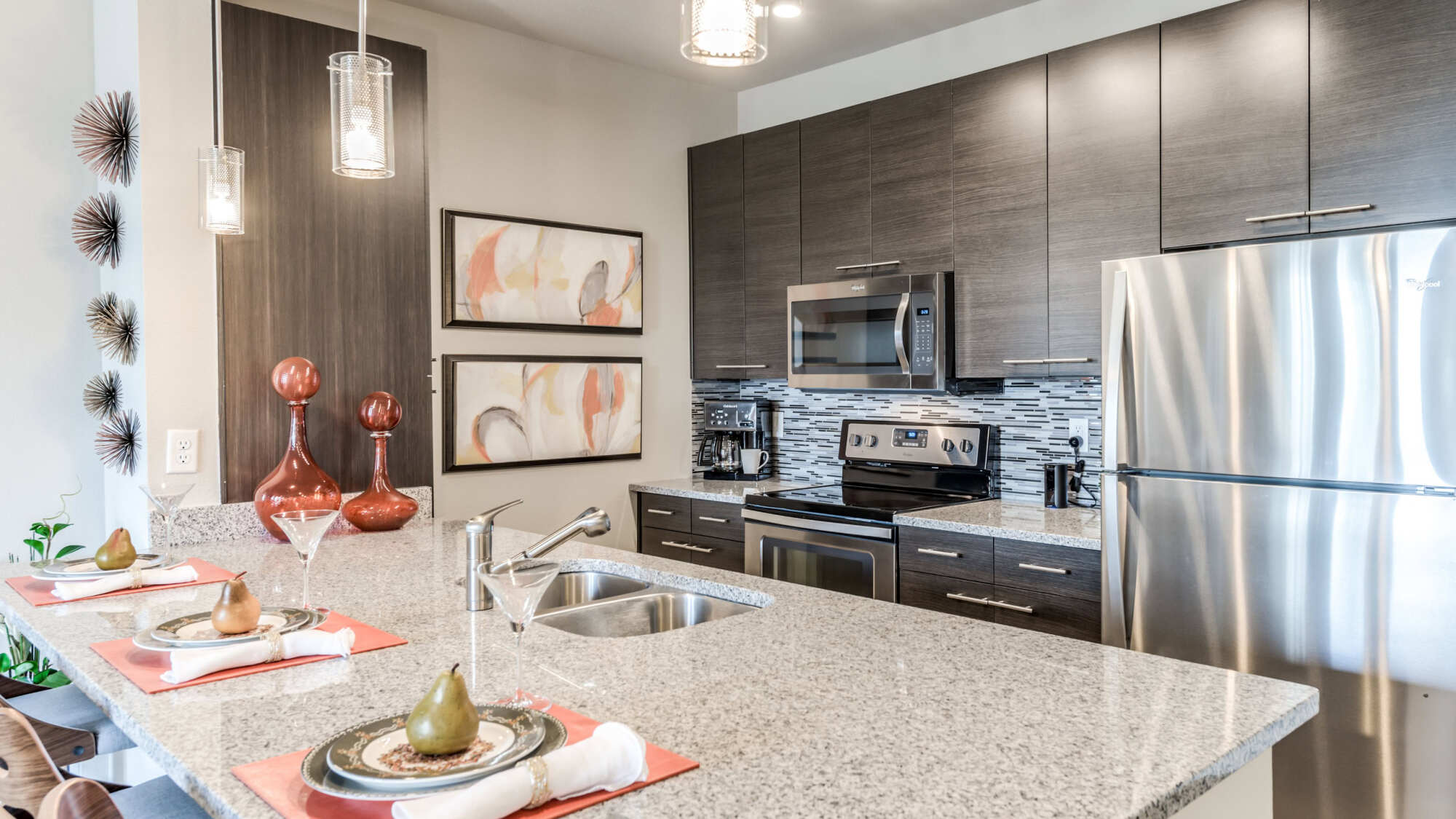 Modern apartment kitchen with dark wood cabinets, light granite countertops, a glass-tile backsplash, stainless steel appliances, pendant lighting, and a staged breakfast bar.