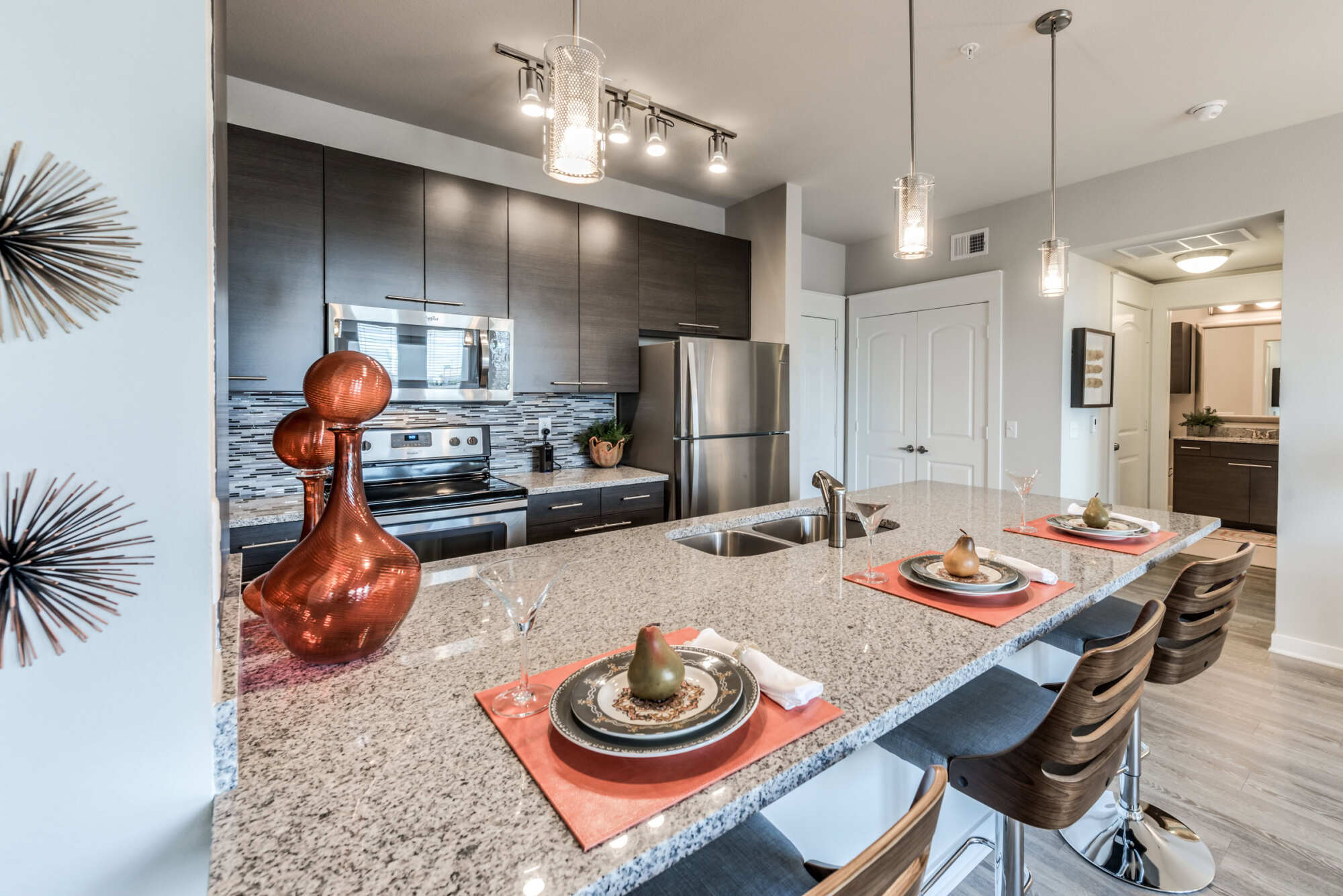 Apartment kitchen with a granite island and bar stools, dark wood cabinetry, glass-tile backsplash, stainless steel appliances, and pendant lights over wood-look flooring.
