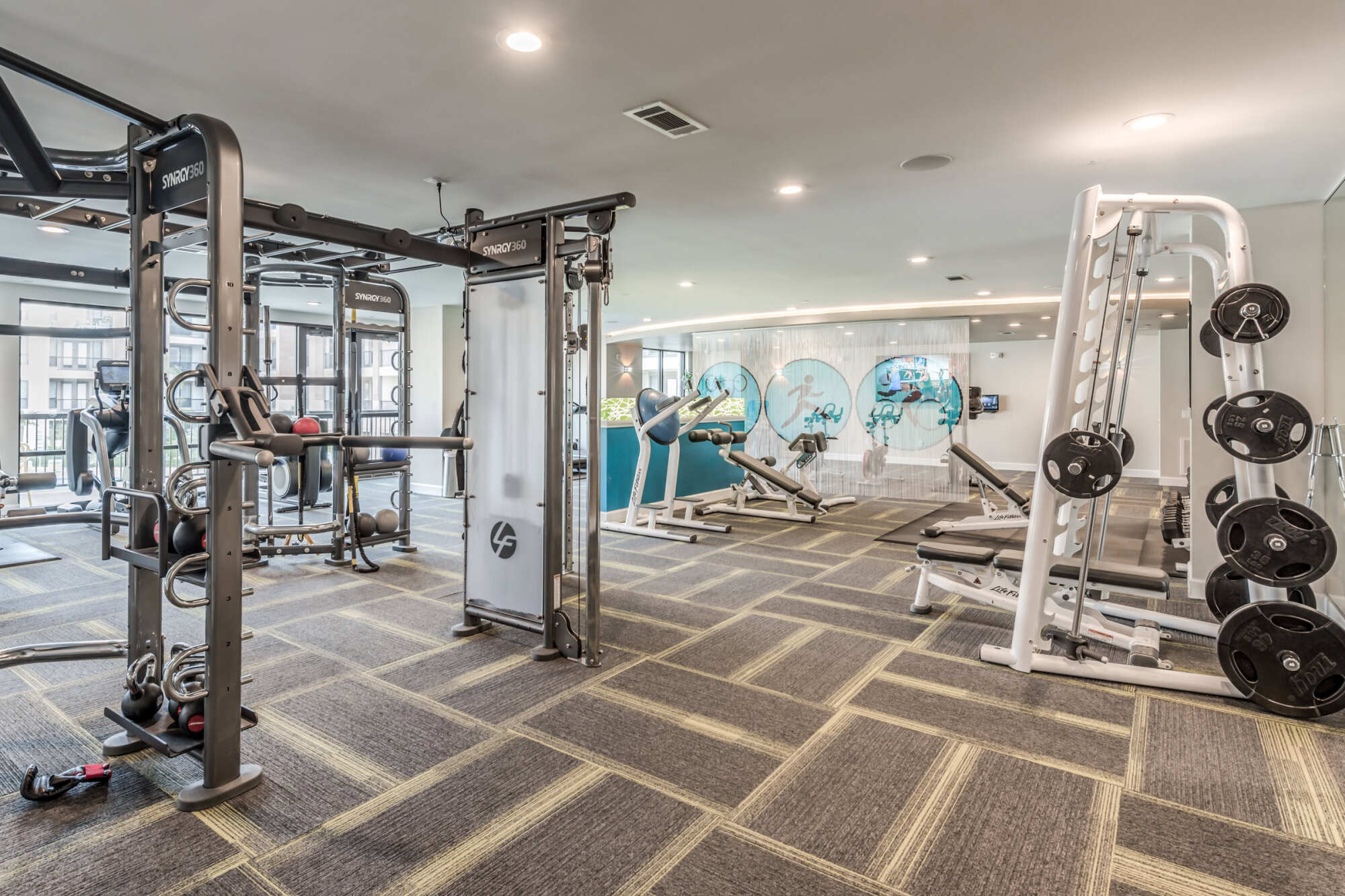 Strength-training section of the fitness center with a Synrgy training rig, weight plates on a rack, adjustable benches, and motivational graphics on the wall.