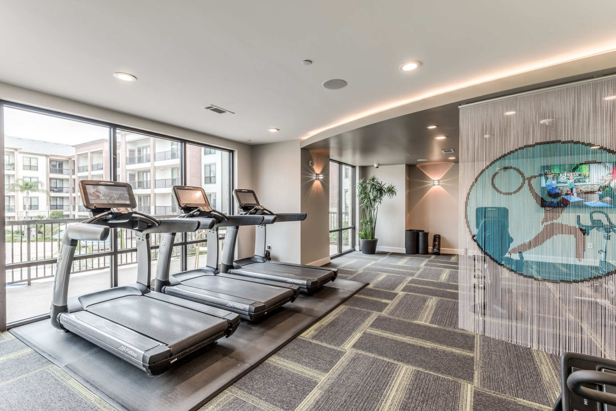 Gym cardio area with a row of treadmills facing large windows that overlook the courtyard, a potted plant, and a beaded-curtain divider with a wall-mounted TV.