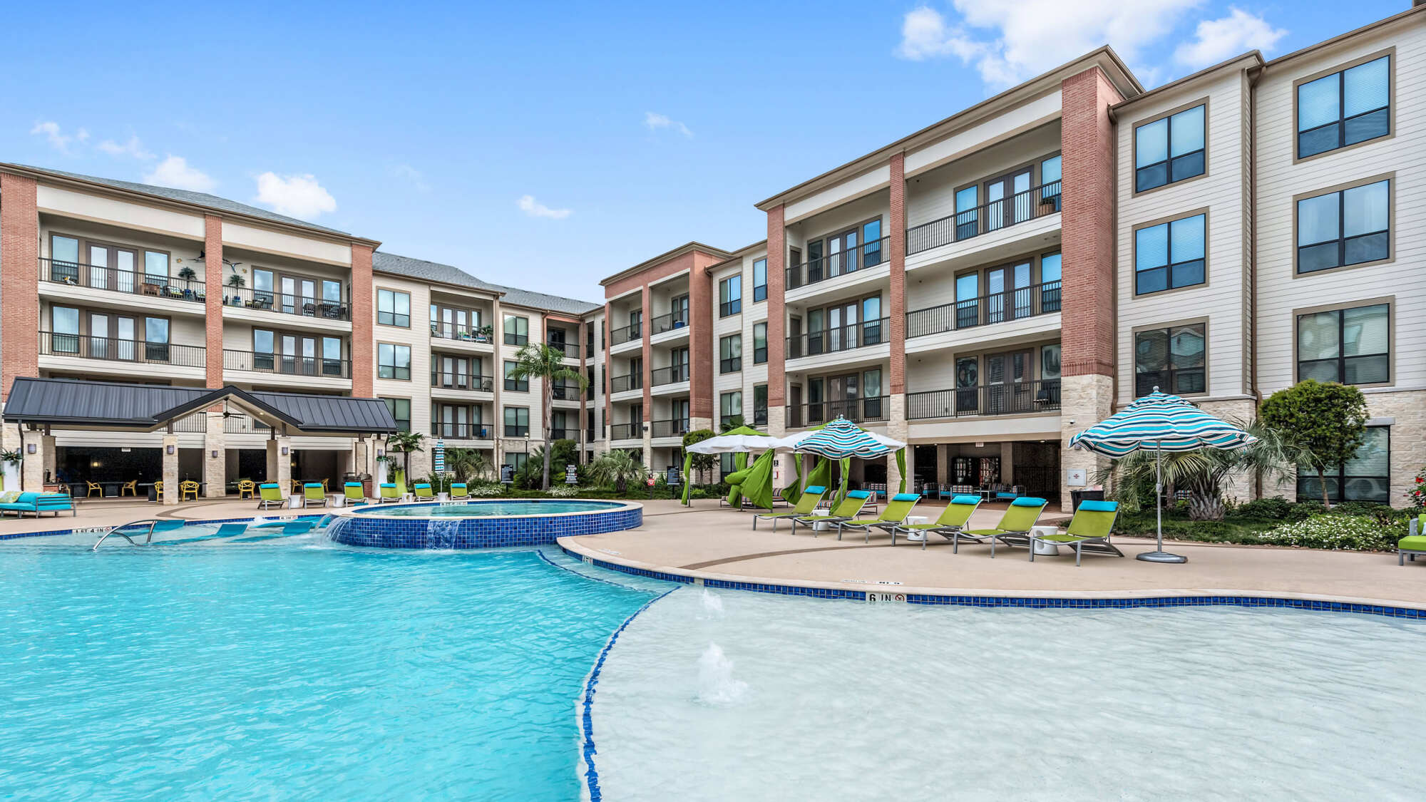 Resort-style courtyard pool with a tanning ledge and bubblers, surrounded by lounge chairs, striped umbrellas, a cabana, and multi-story apartment buildings.