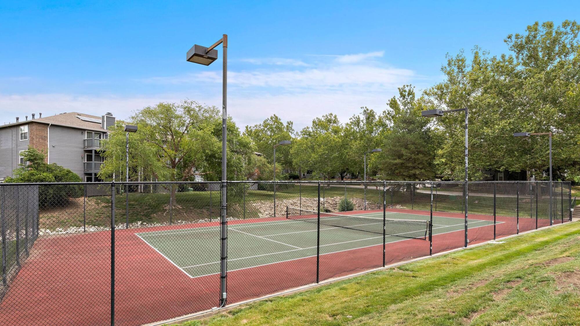 Fenced tennis court at Timberlane Village Apartments in Kansas City, MO, framed by mature trees under a clear blue sky.