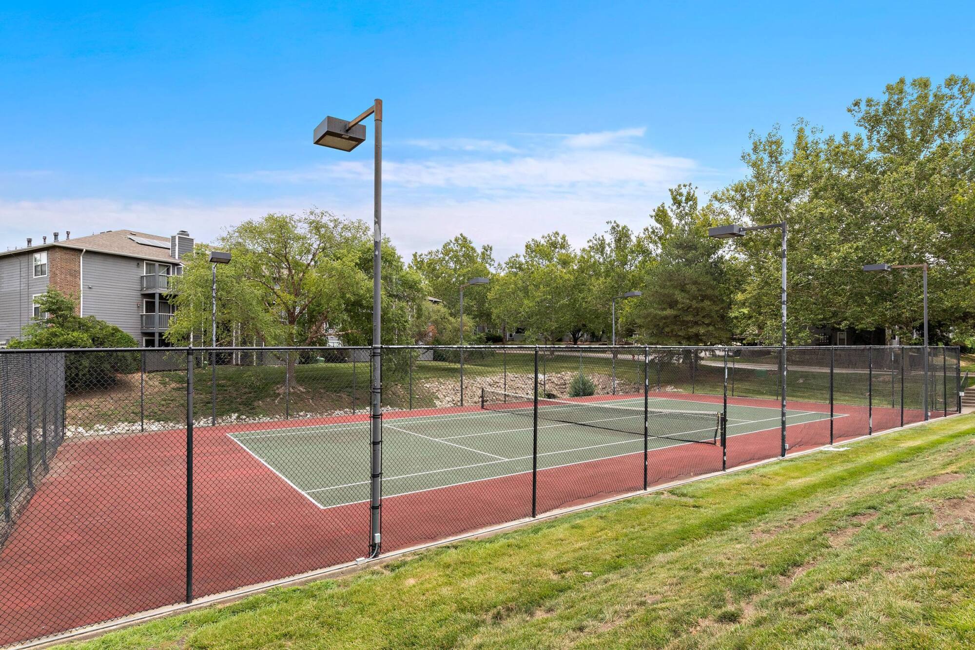 Lighted tennis court at Timberlane Village in Kansas City, MO with a grassy lawn and shade trees along the fence line.