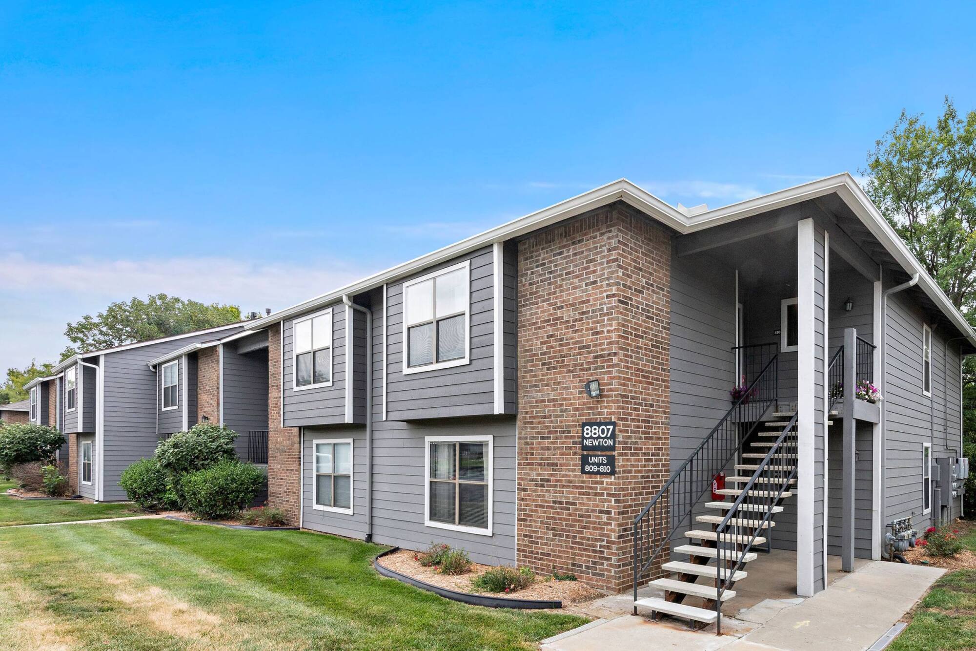 Two-story brick and grey siding apartment building at Timberlane Village in Kansas City, MO with a landscaped lawn and stairway.