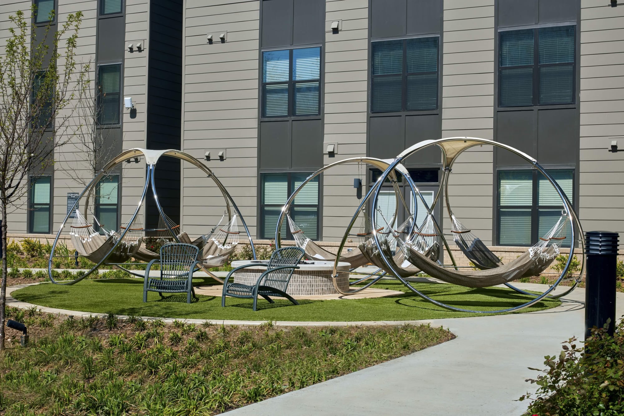 Courtyard lawn with sculptural stainless-steel hammock loungers and metal chairs in front of the building's gray façade.