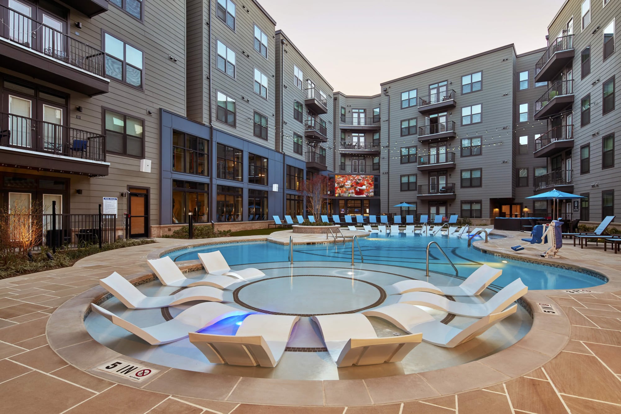 Resort-style courtyard pool at dusk with in-water chaise loungers, a sun shelf, string lighting, and the illuminated apartment building behind it.