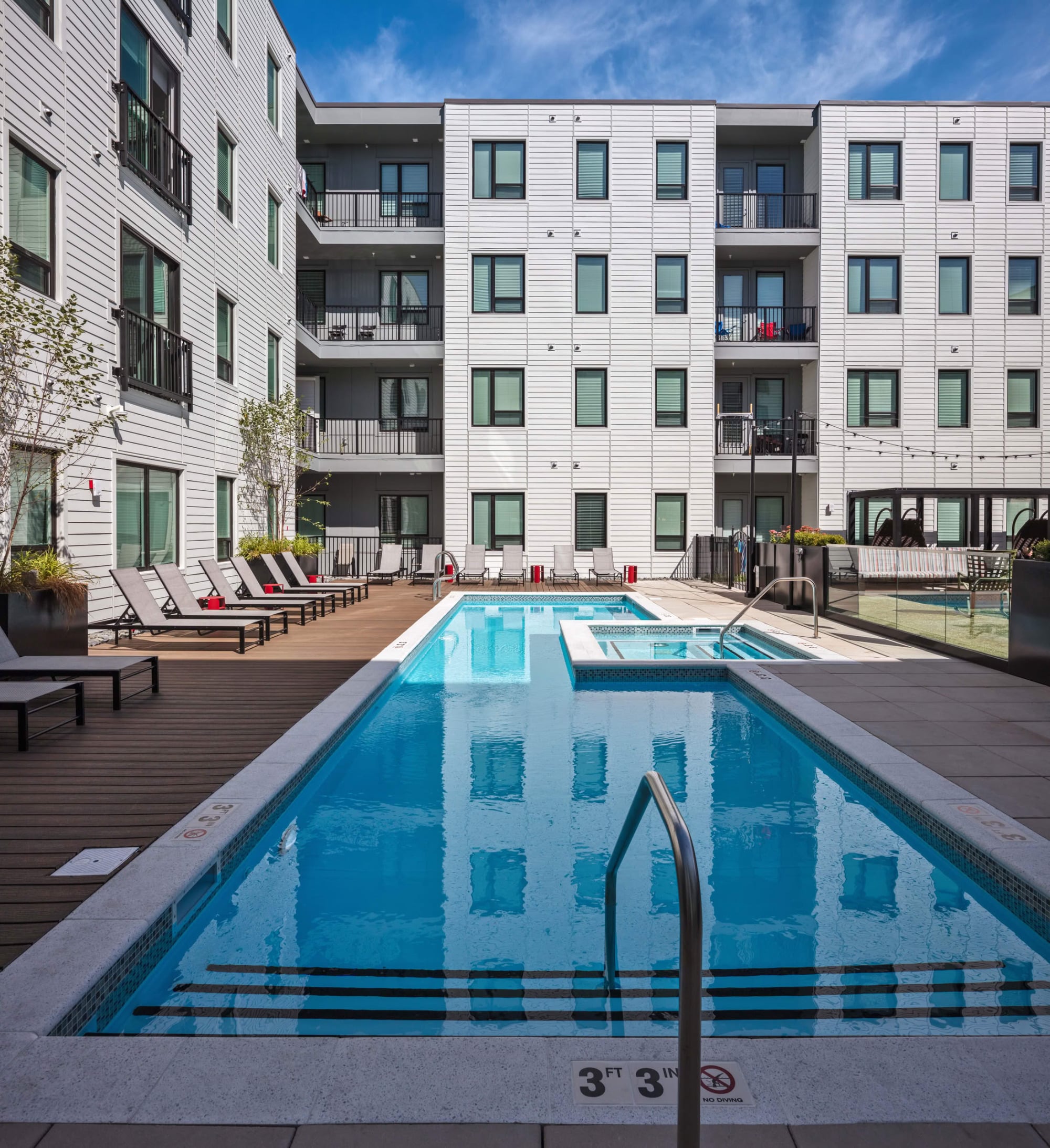 Courtyard swimming pool and spa with chaise loungers and red side tables on a wood-look deck, surrounded by the white balconied building under a blue sky.