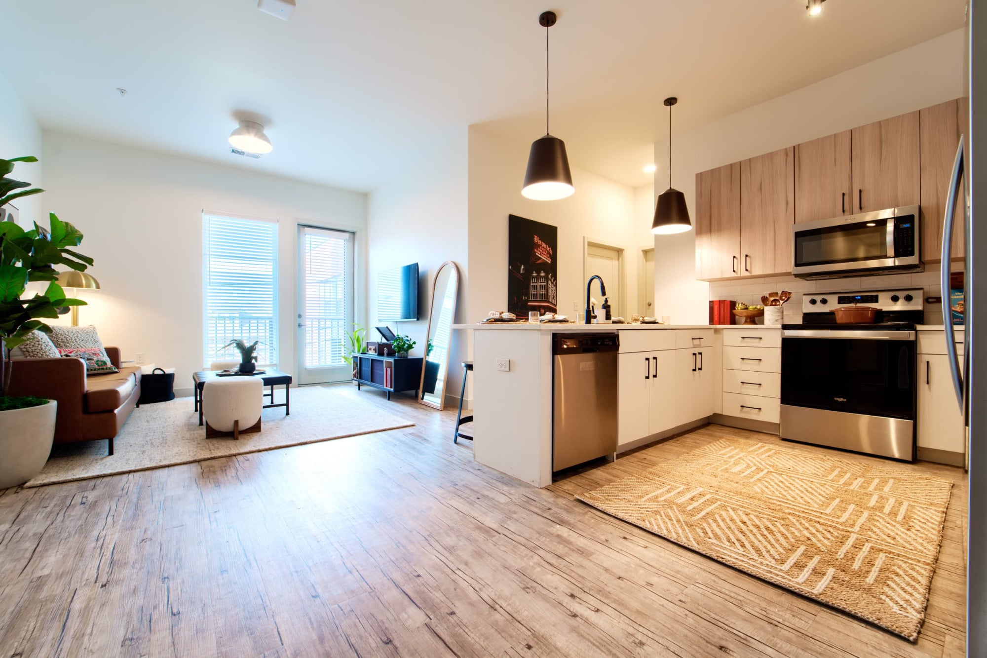Open-concept apartment with a living area and kitchen, featuring a leather armchair, light wood cabinets, white island, stainless steel appliances, and a patterned rug.