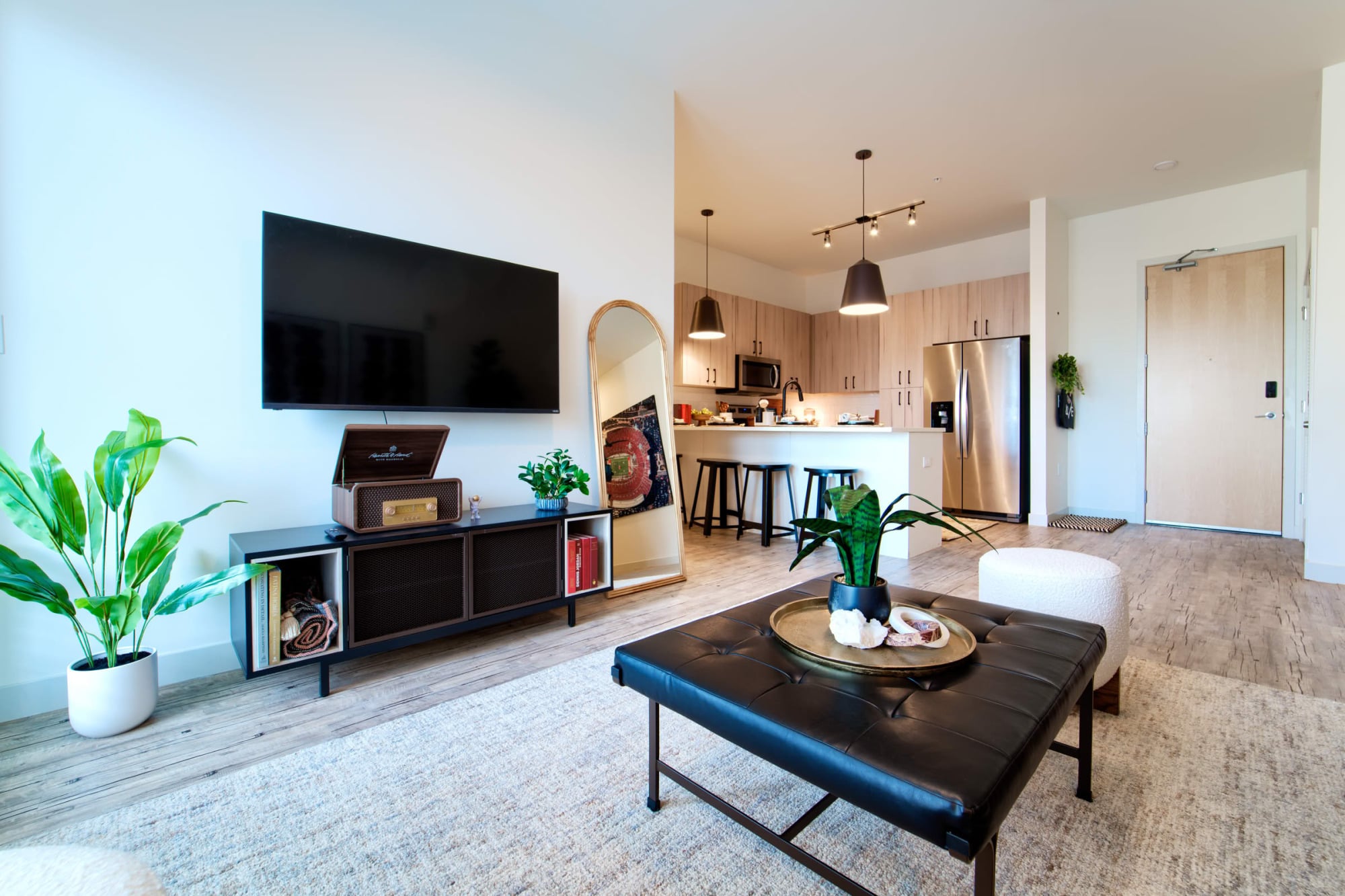 Open living and kitchen area with a wall-mounted TV, leaning mirror, black coffee table, potted plants, and a kitchen with bar seating in the background.