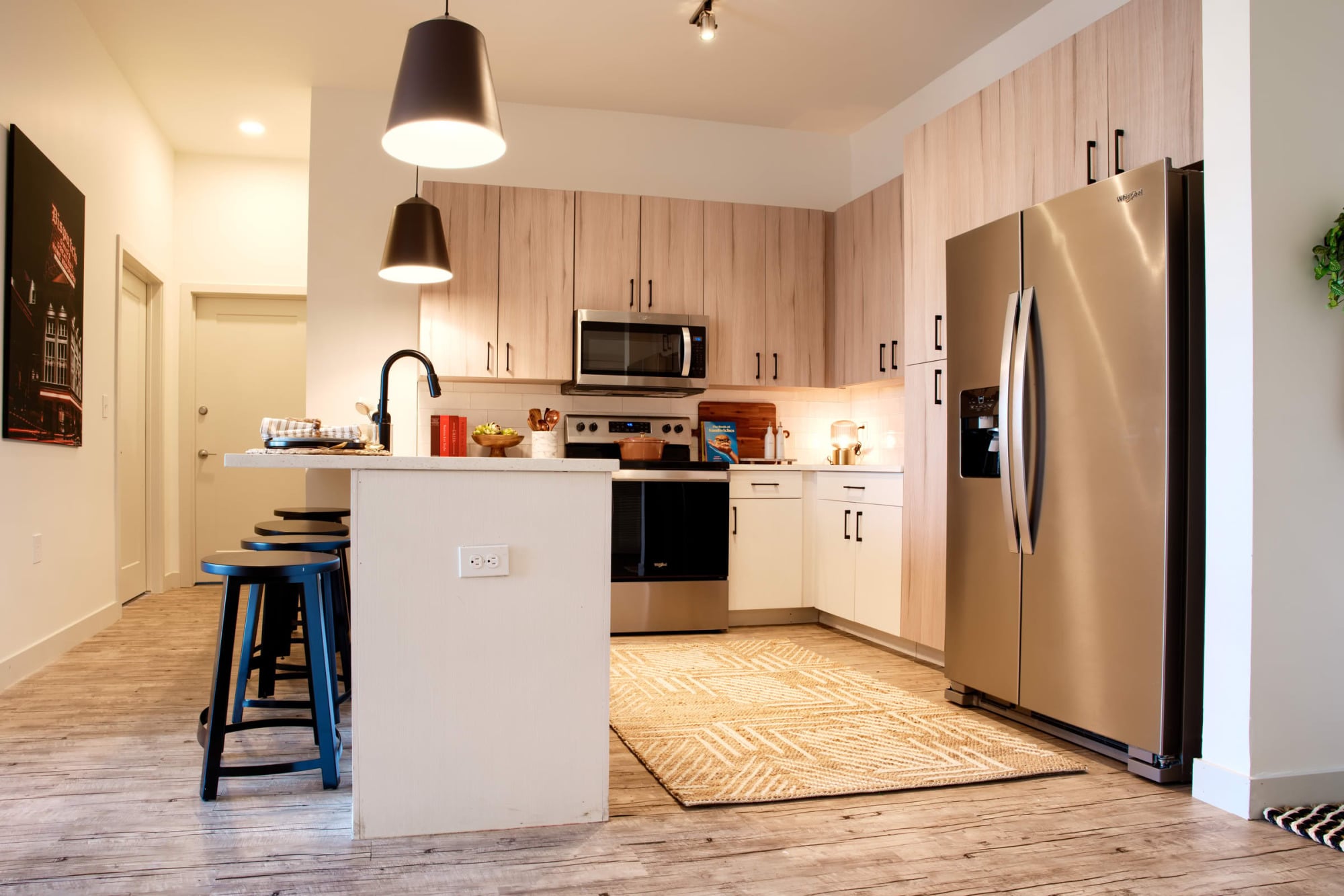 Apartment kitchen with light wood cabinets, a white island with blue bar stools, stainless steel appliances, pendant lights, and a patterned rug.