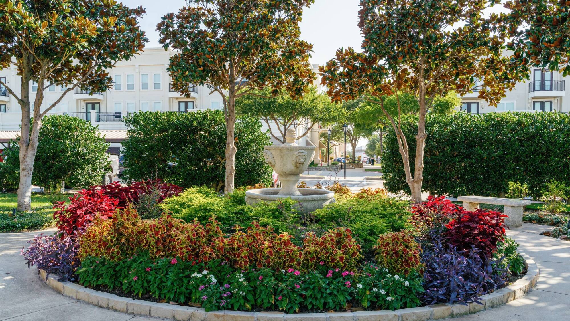 Circular garden with colorful plants, a stone fountain in the center, and trees in front of a building.