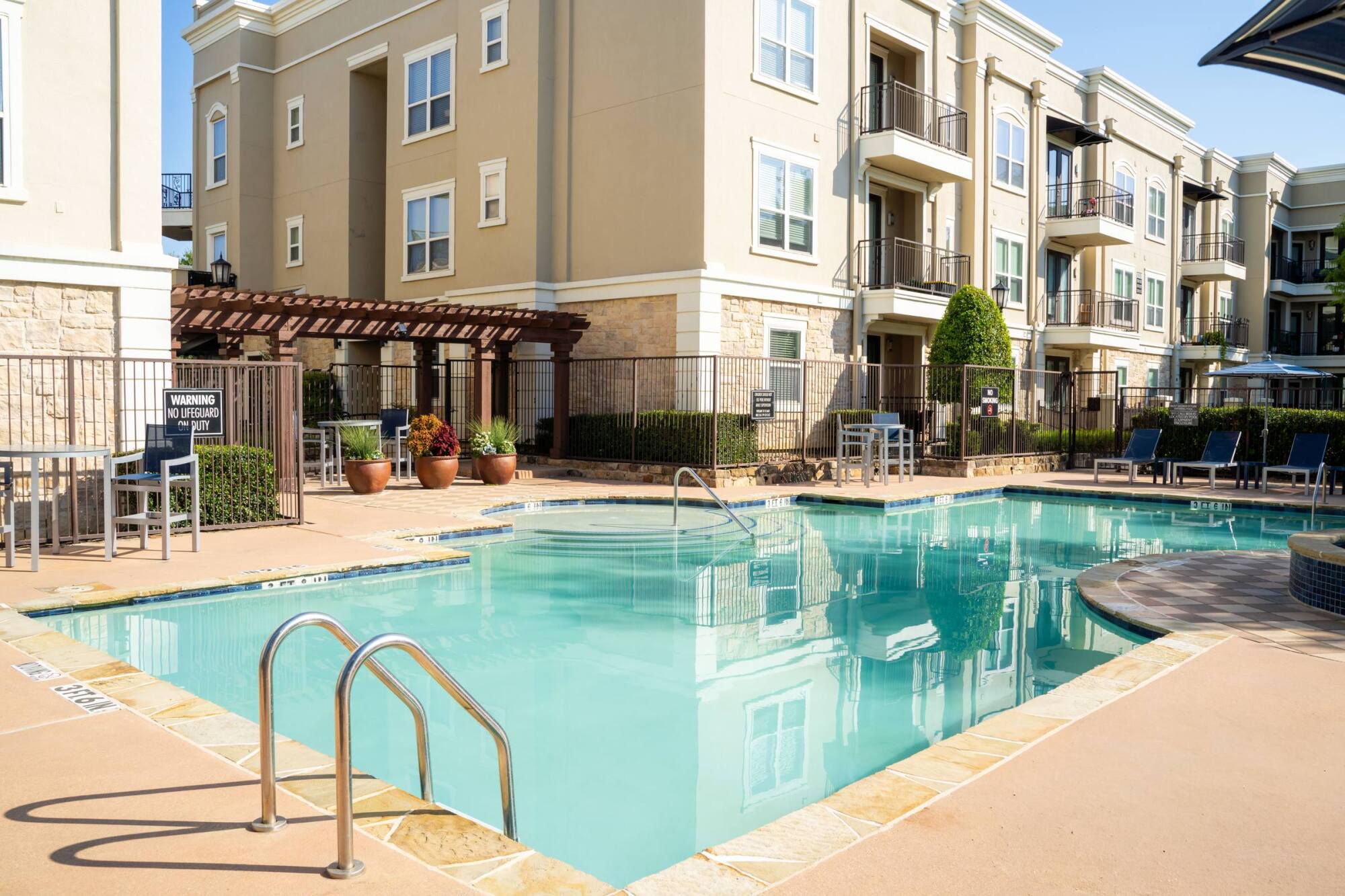 Outdoor apartment pool with clear water, surrounded by lounge chairs and beige multi-story buildings.