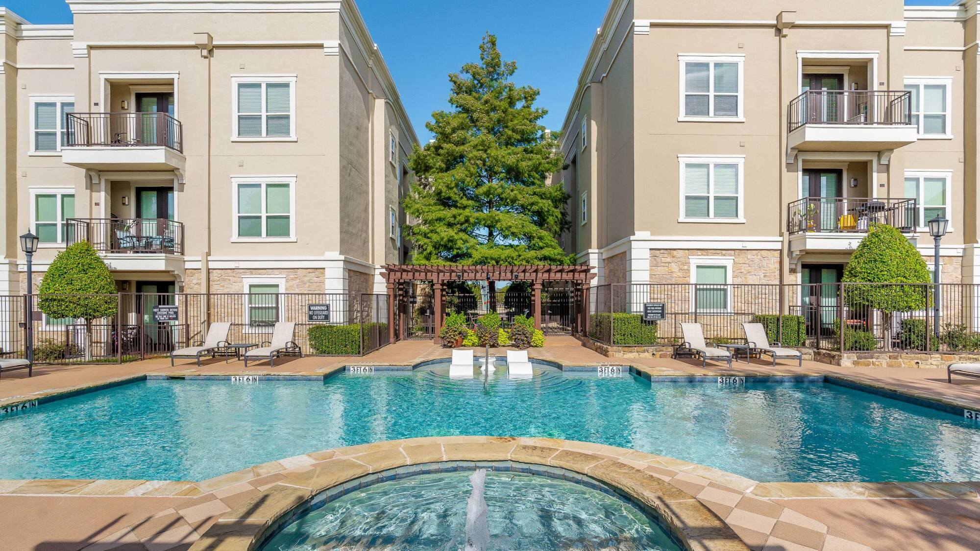 Outdoor pool with hot tub, lounge chairs, and two beige apartment buildings on either side, under a clear sky.