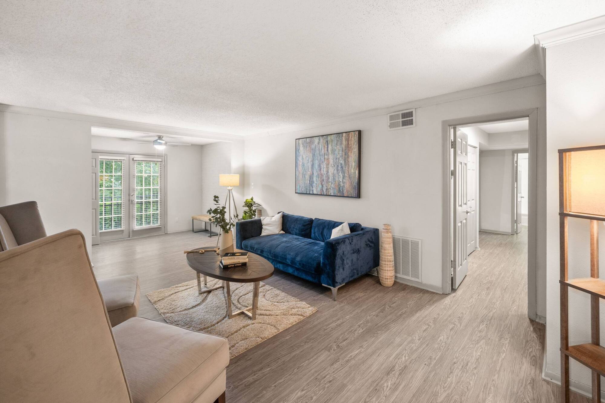 Living room at The Whitney with wood-plank vinyl flooring, a blue velvet sofa, and patio doors letting in natural light.