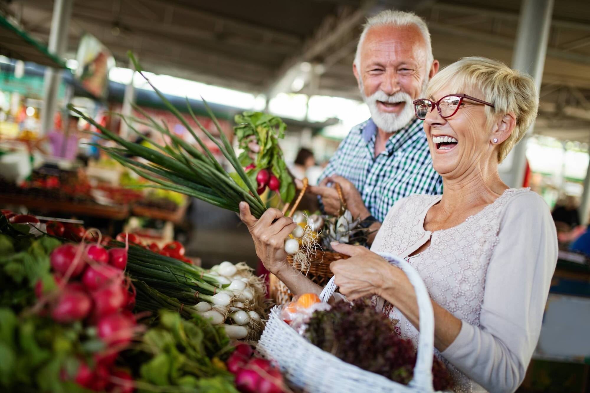 Smiling older couple shopping for fresh vegetables at an outdoor market, holding produce and a basket.