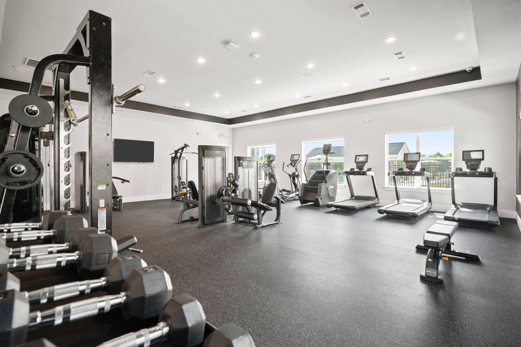 Modern gym with dumbbells, weight machines, treadmills, and elliptical machines, in a well-lit room with windows.