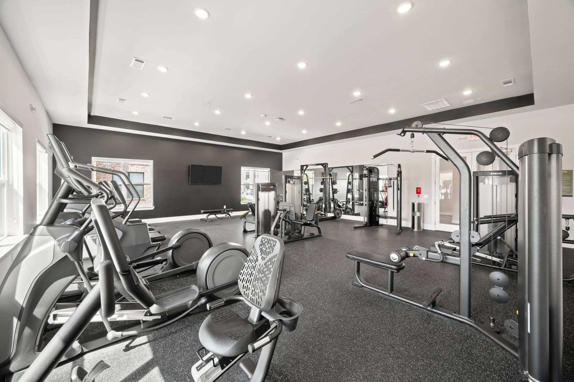 Modern gym with cardio machines, weight machines, and benches in a bright room with large windows.