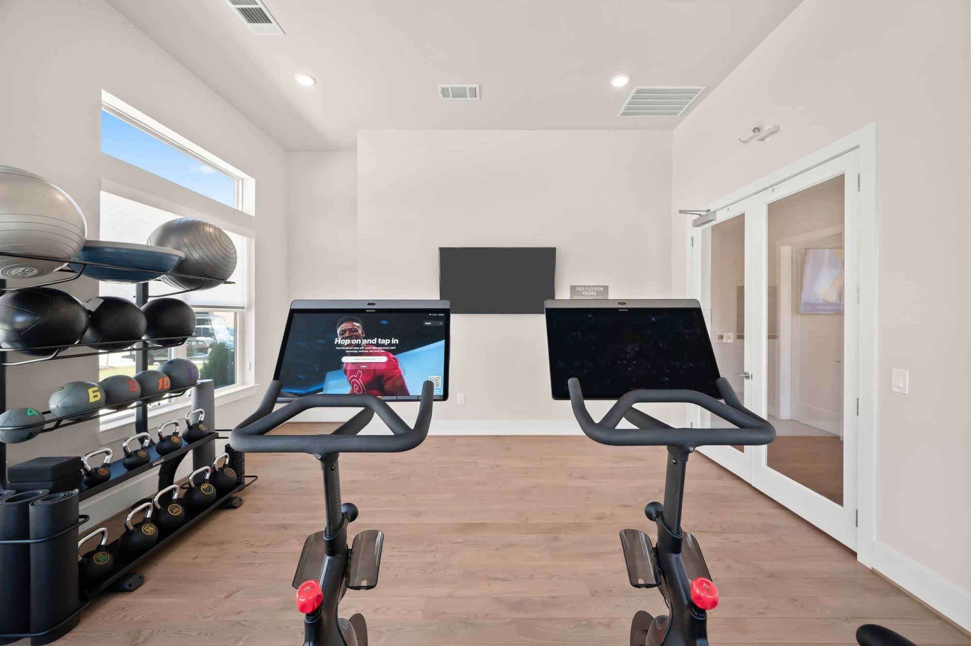 Two stationary bikes face a TV in a bright home gym with weights and exercise balls on shelves.