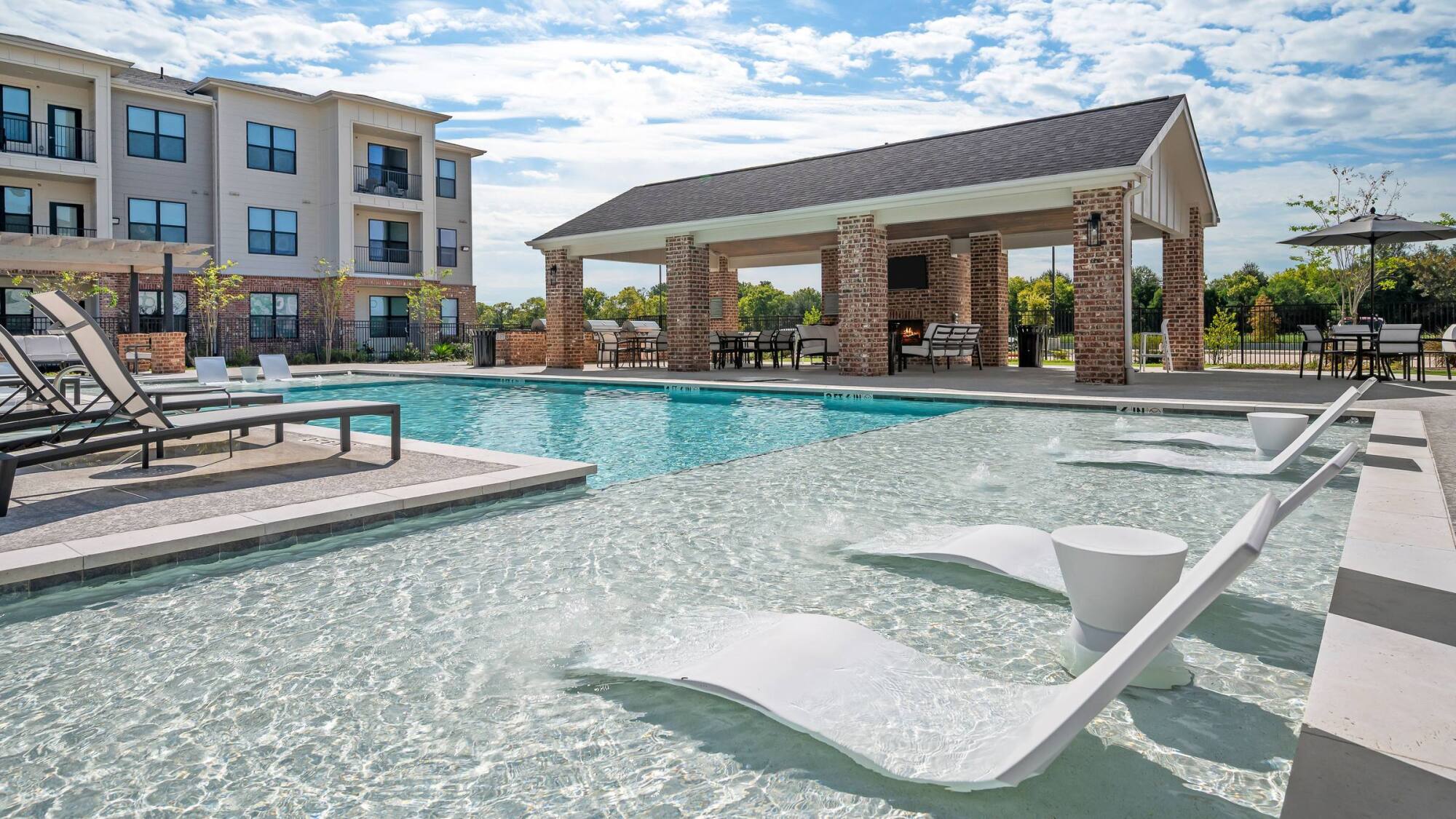 Modern outdoor pool with in-water lounge chairs, shaded patio area, and apartment building in the background.