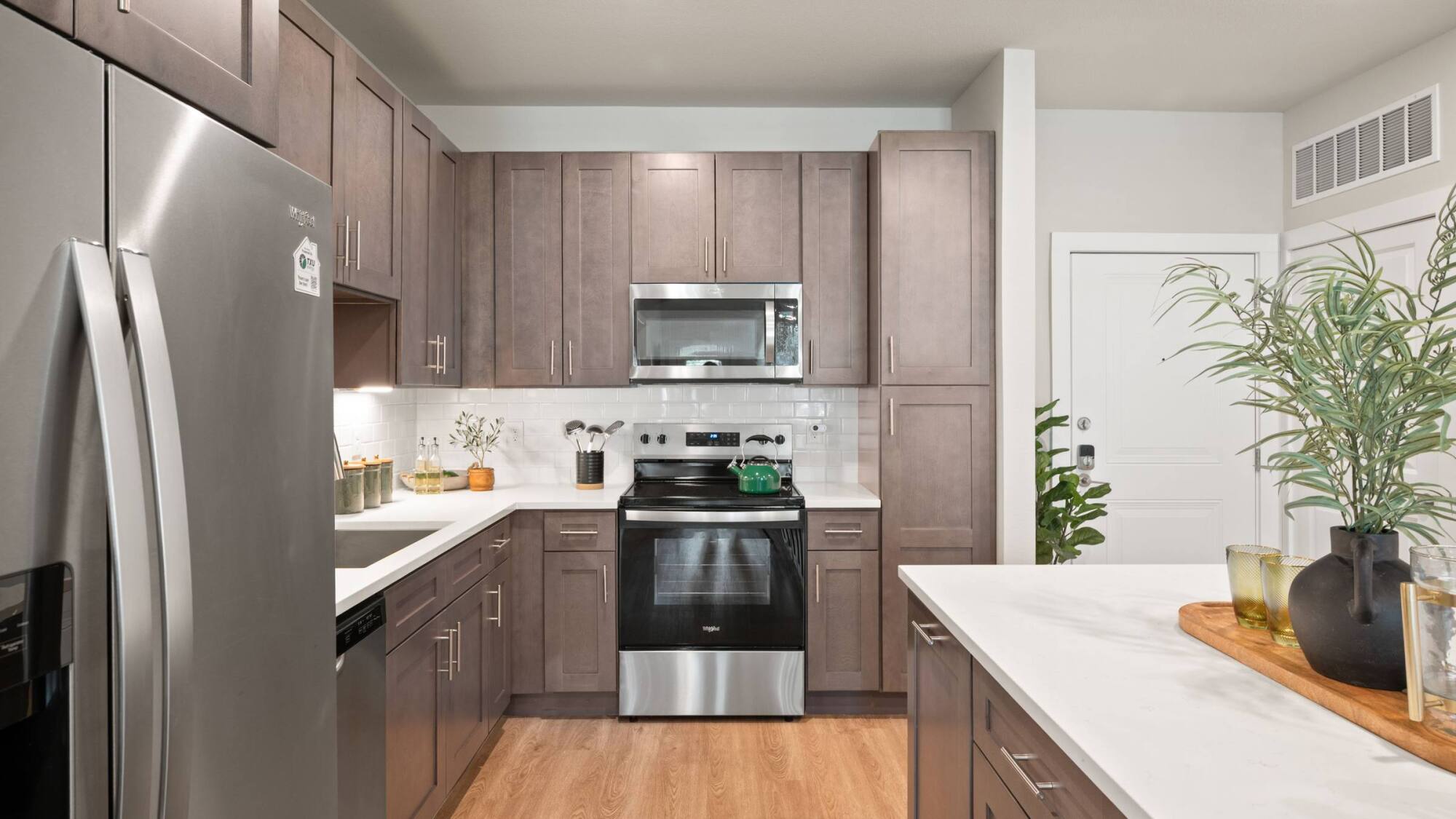 Modern kitchen with stainless steel appliances, brown cabinets, white countertops, and wood flooring.