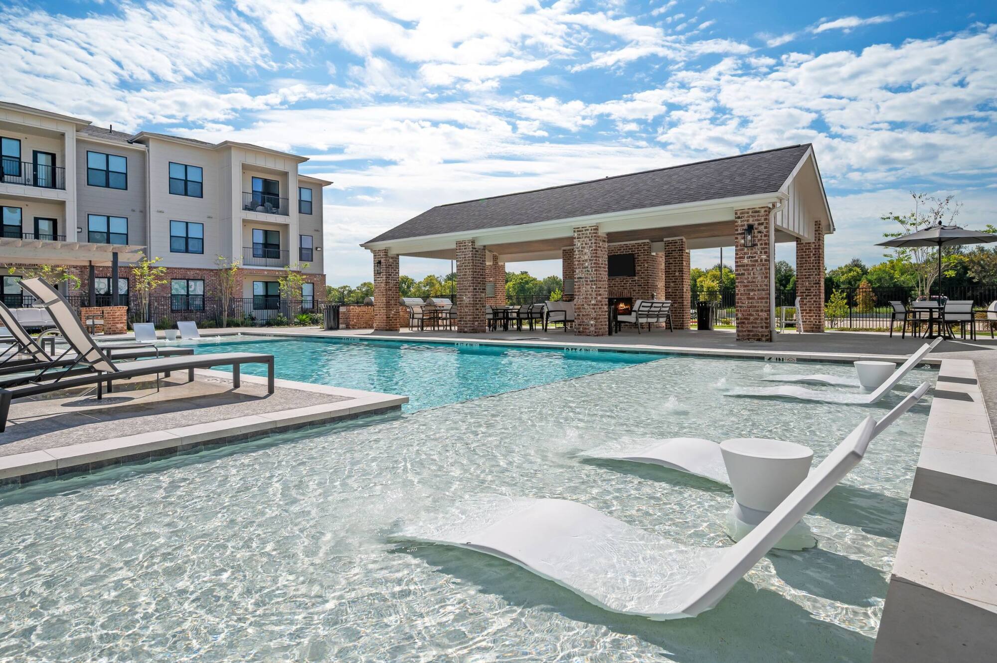 Modern outdoor pool with lounge chairs, shaded pavilion, and apartment building in the background under blue sky.