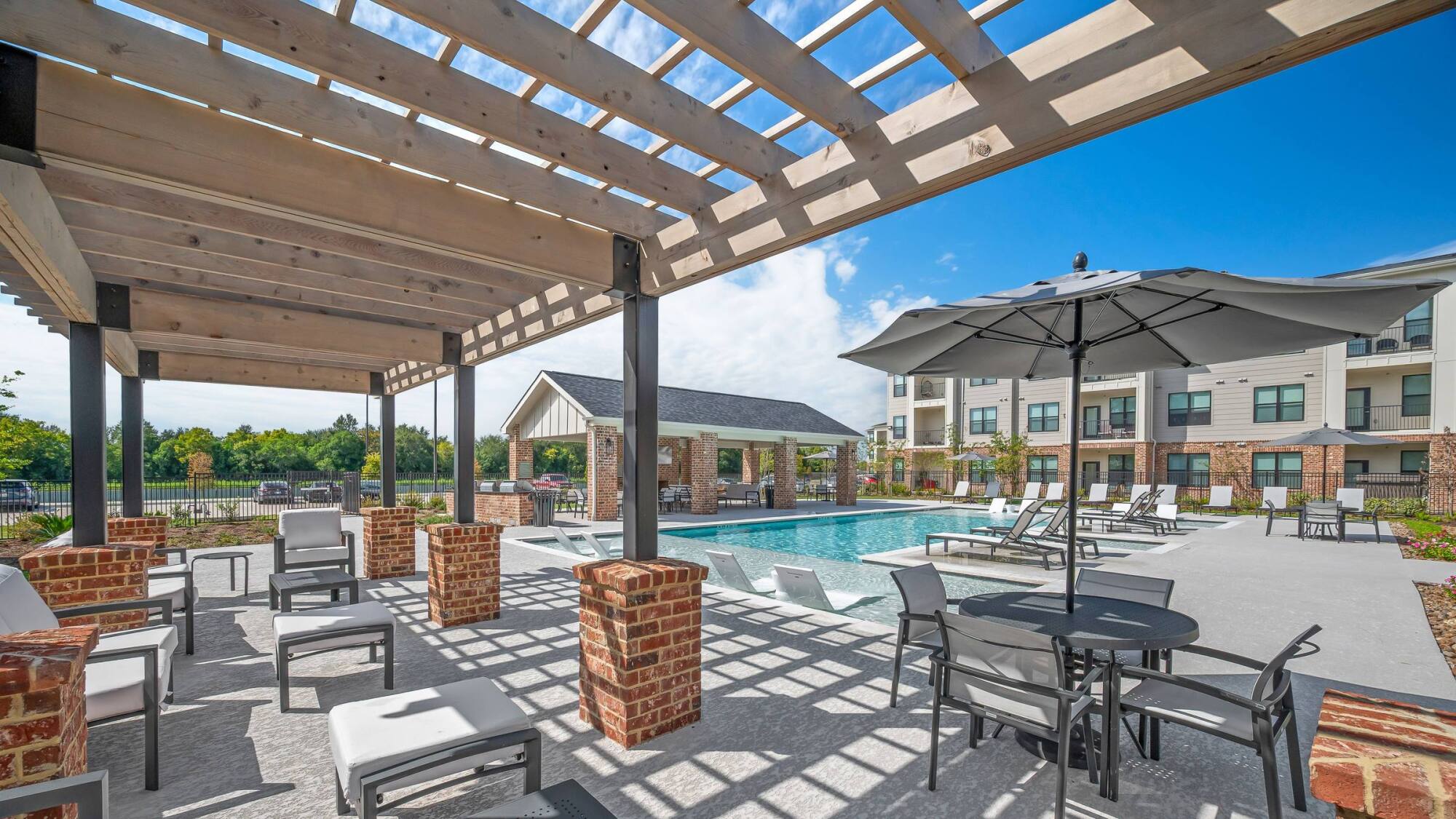 Outdoor pool area with lounge chairs, tables, umbrellas, and a pergola beside an apartment building on a sunny day.