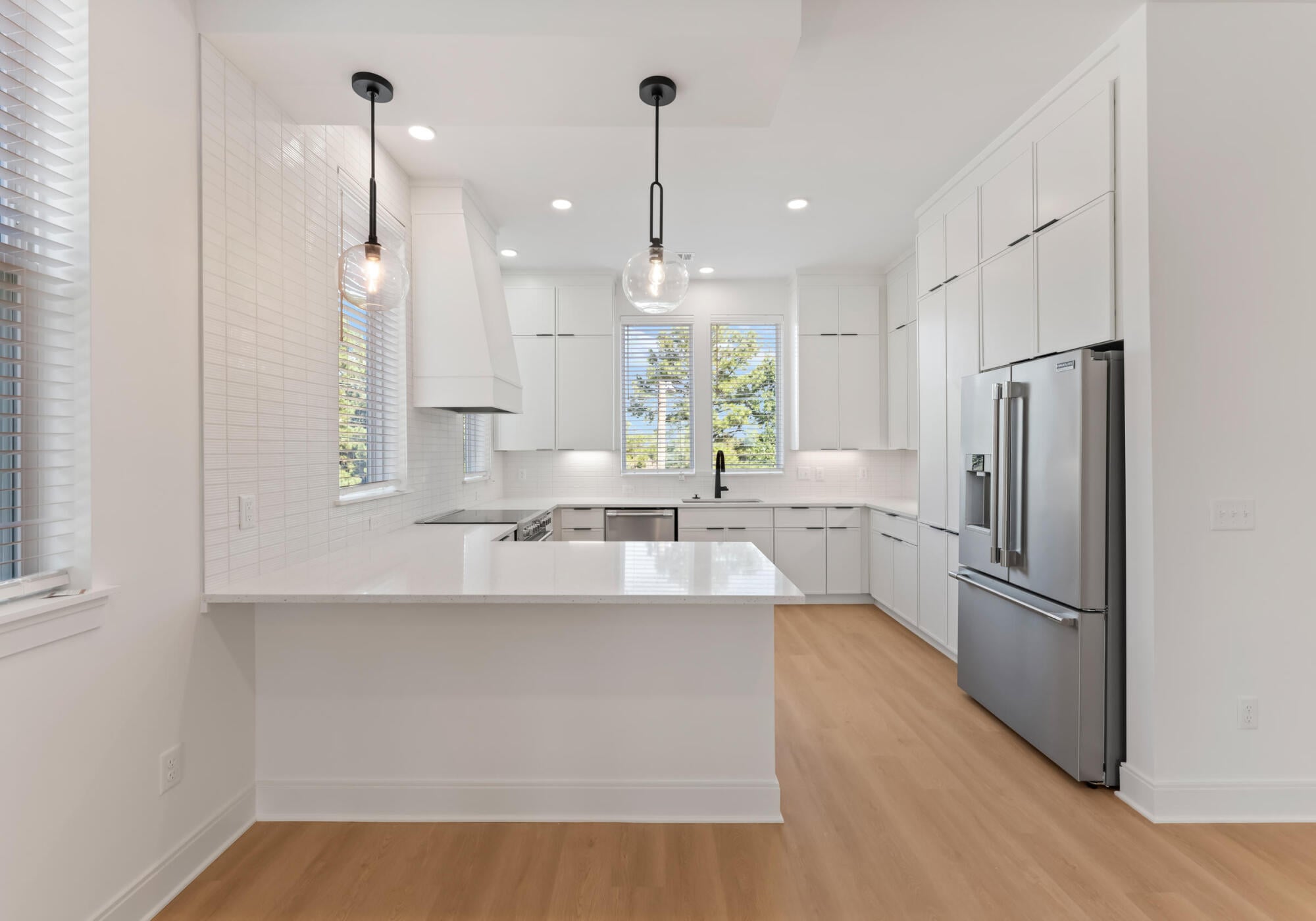 Modern white kitchen with island, pendant lights, stainless steel appliances, and light wood flooring.