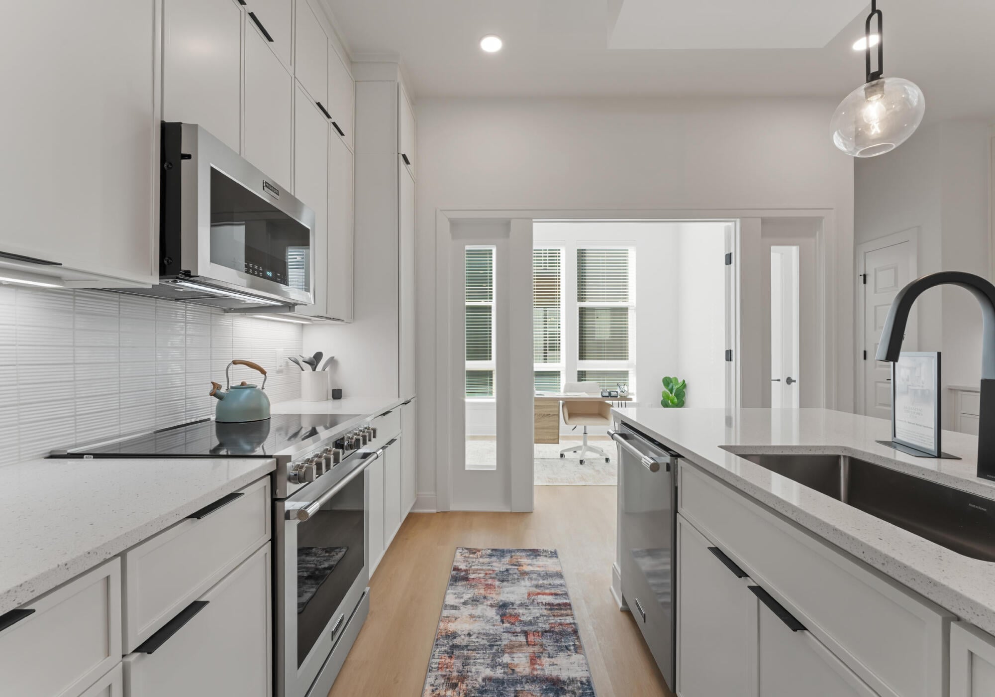 Modern white kitchen with stainless steel appliances, island sink, and a view of a bright office through glass doors.