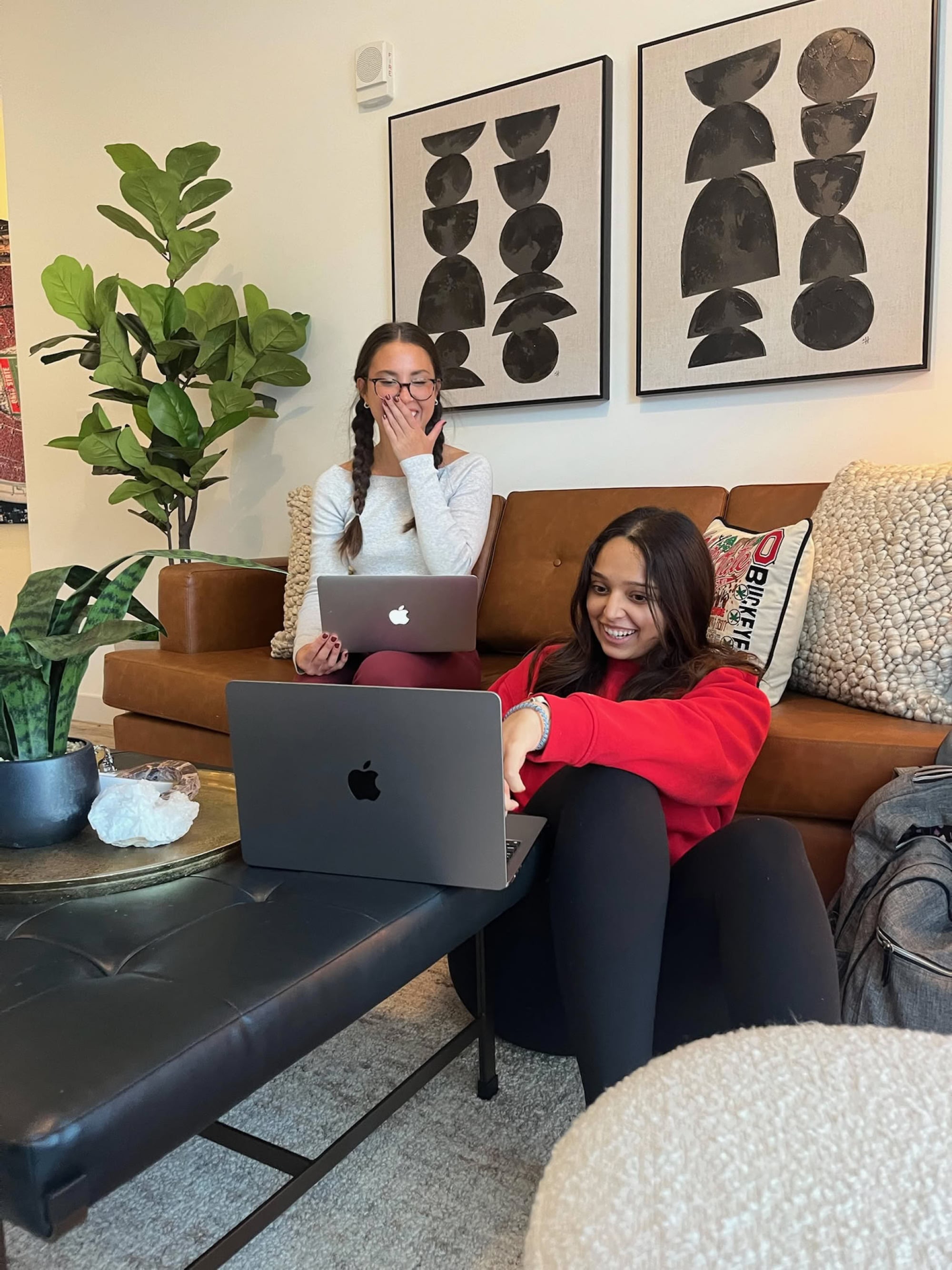 Two young women laughing while using laptops on a leather couch in a cozy, art-filled resident lounge.