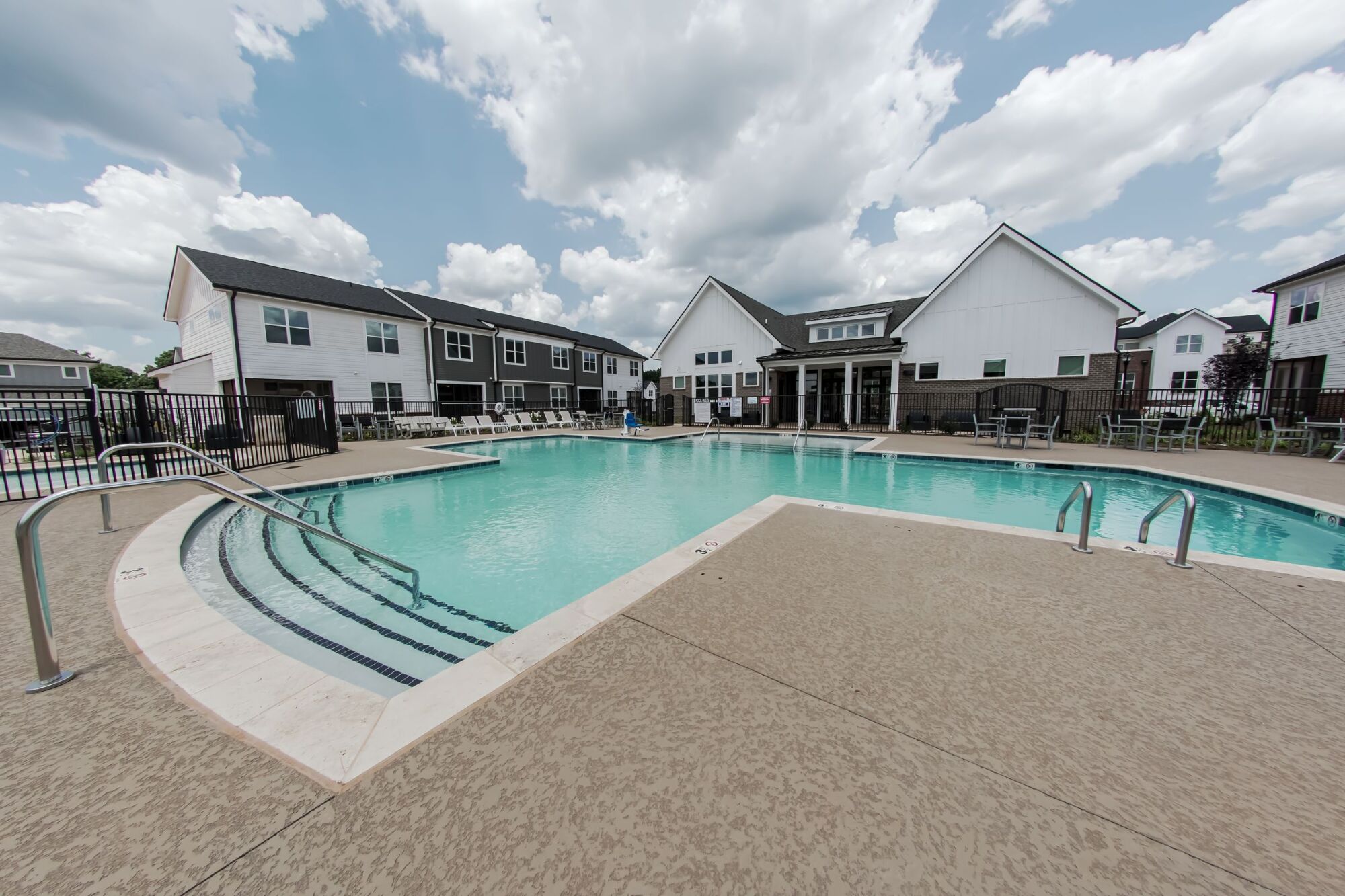 Outdoor swimming pool surrounded by houses and lounging chairs under a partly cloudy sky.