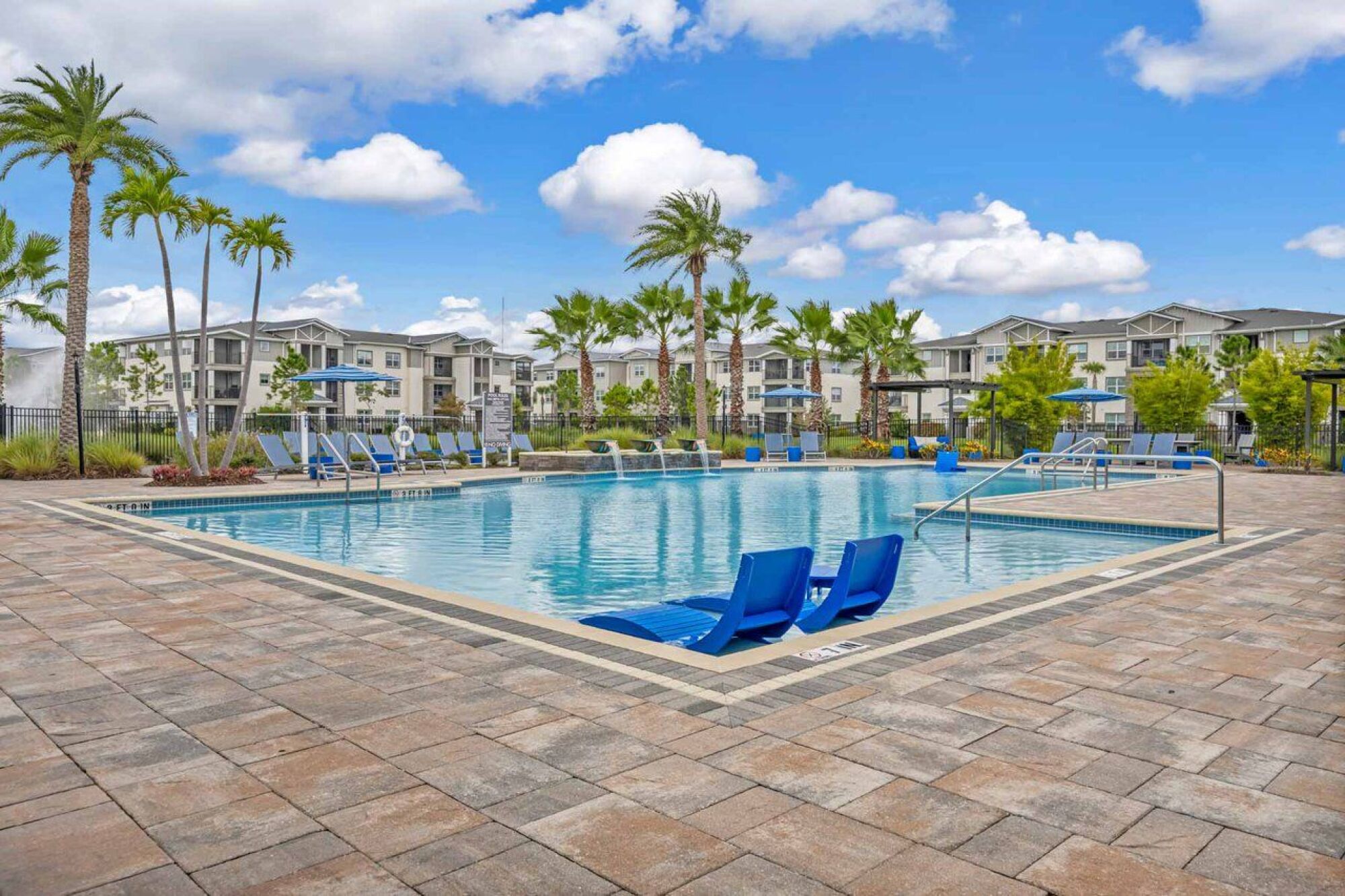 Outdoor swimming pool with blue lounge chairs, palm trees, and modern apartment buildings in the background at Luminary at 95 apartments.