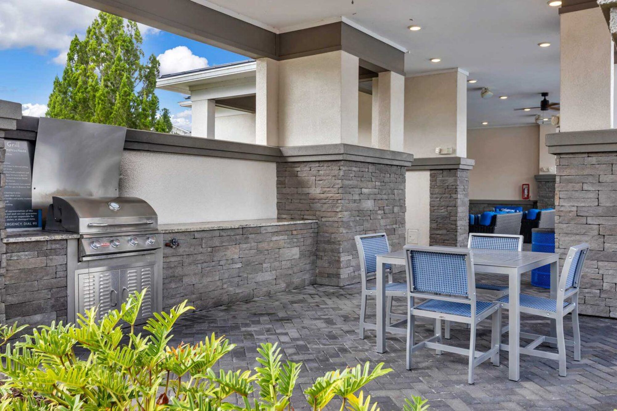 Outdoor patio with a stainless steel grill, stone walls, and a table with blue chairs on a paved floor at Luminary at 95 apartments.