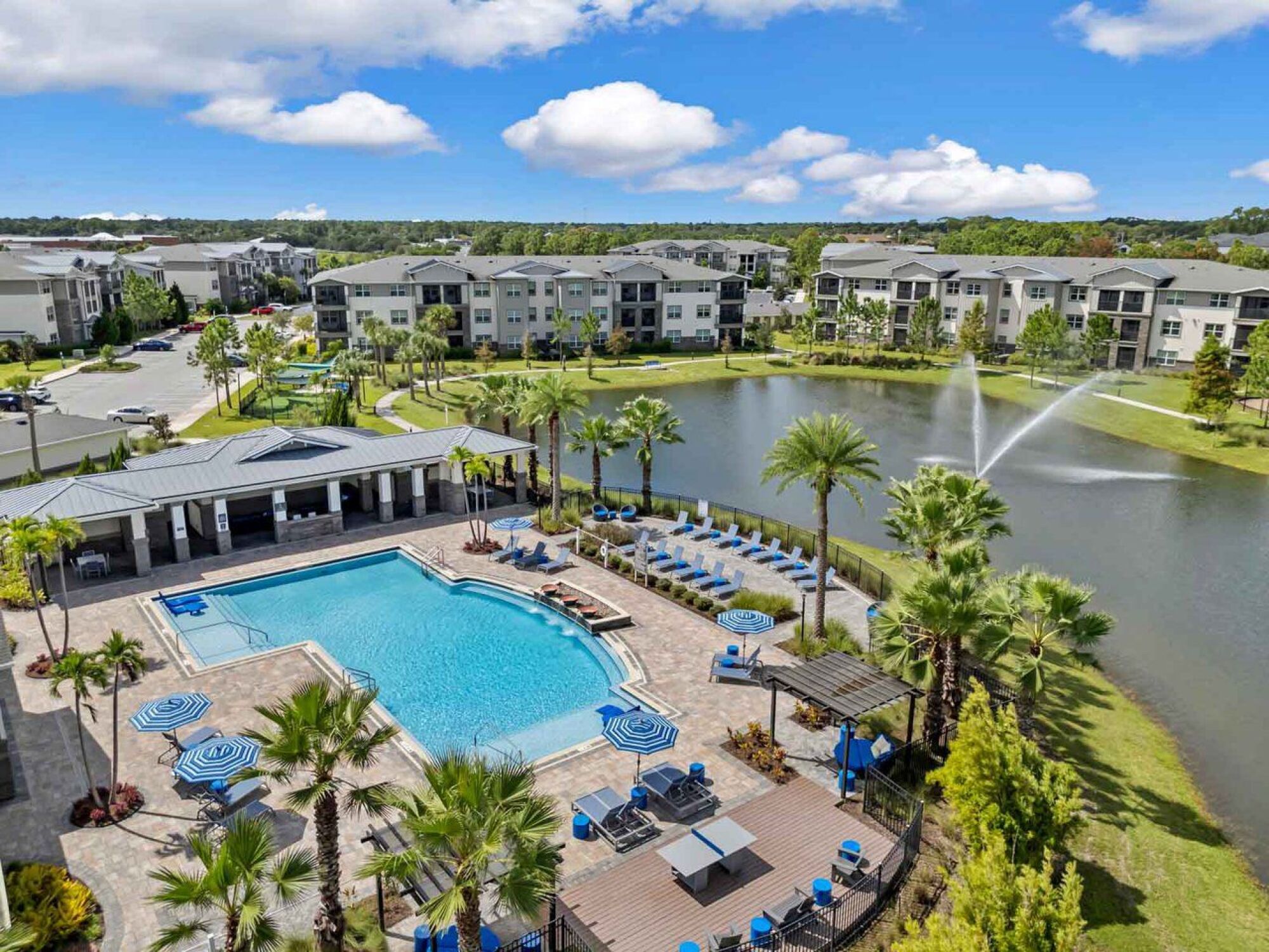 Aerial view of an apartment complex with a pool, lounge chairs, palm trees, pond, and water fountain at Luminary at 95 apartments.