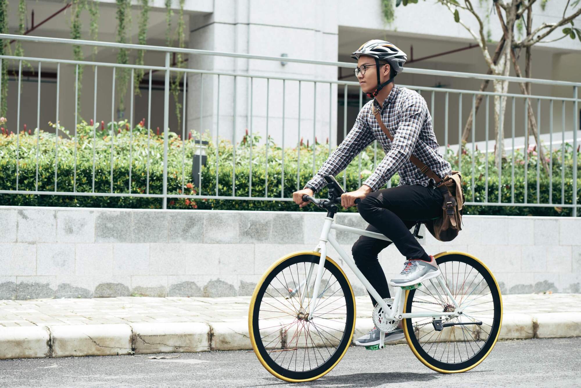 Man wearing a helmet and glasses riding a bicycle on a city street, carrying a brown shoulder bag.