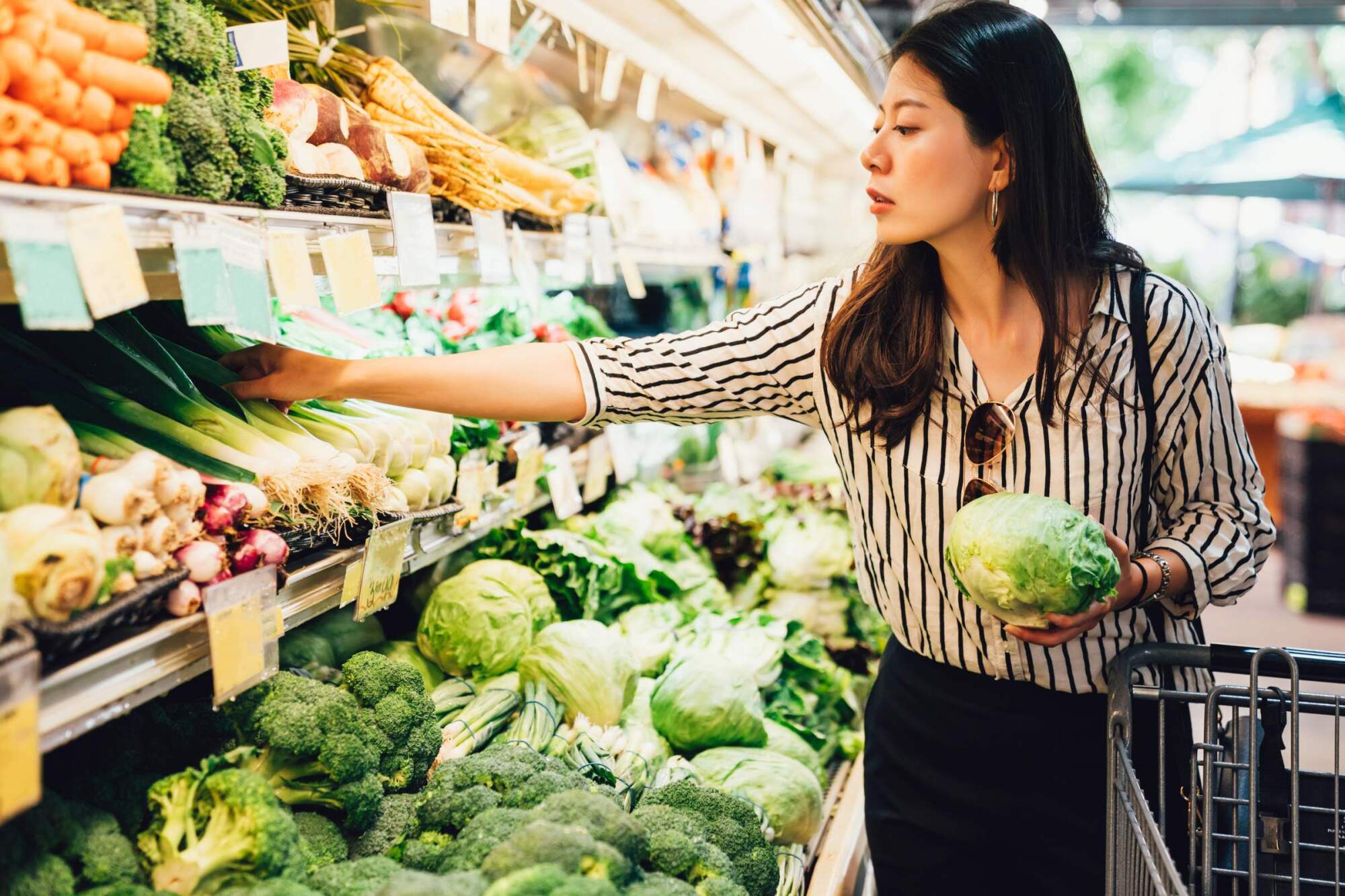 Woman shopping for fresh vegetables, holding a cabbage and reaching for leeks in a grocery store produce aisle.