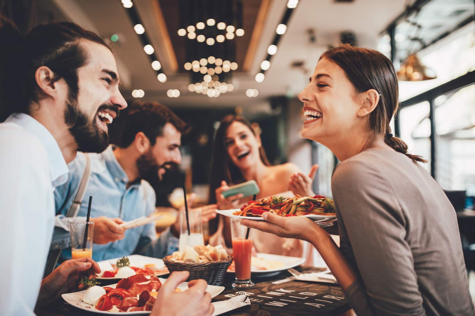 Four friends laughing and enjoying food and drinks together at a lively restaurant table.