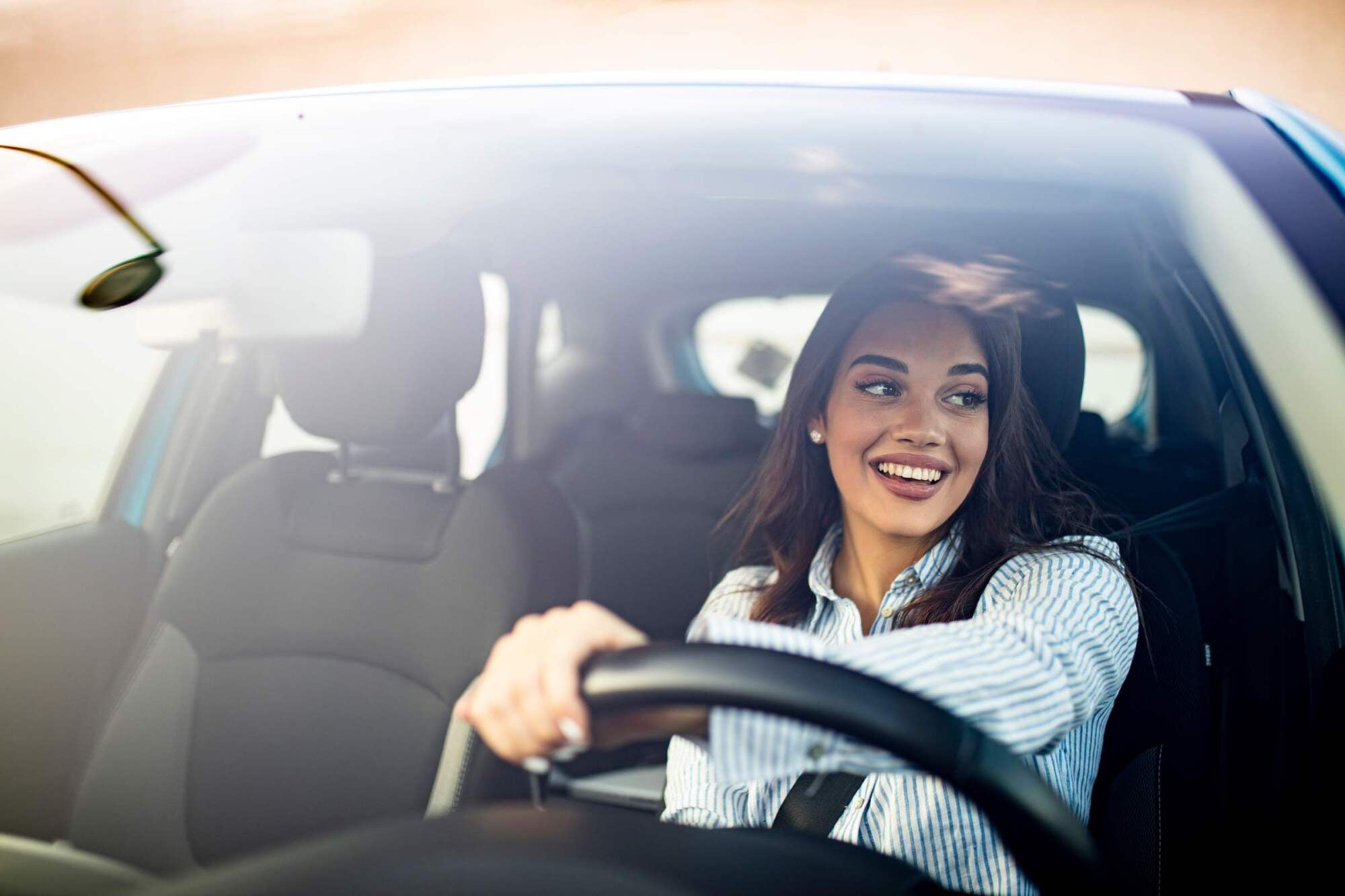 Woman smiling while driving a car, looking out the window with one hand on the steering wheel.