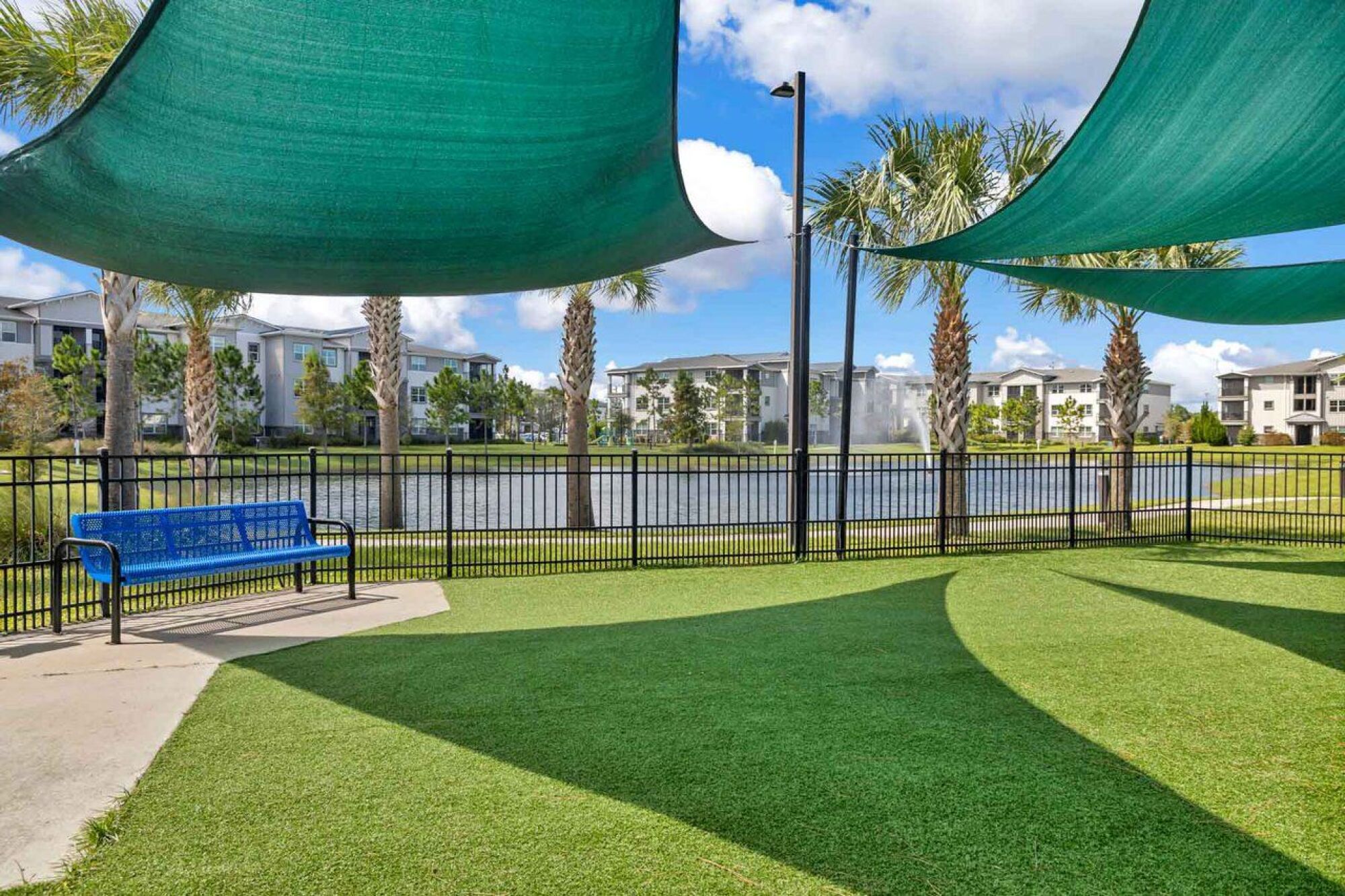 Shaded grassy area with palm trees, a blue bench, and a pond with fountain in the background at Luminary at 95.
