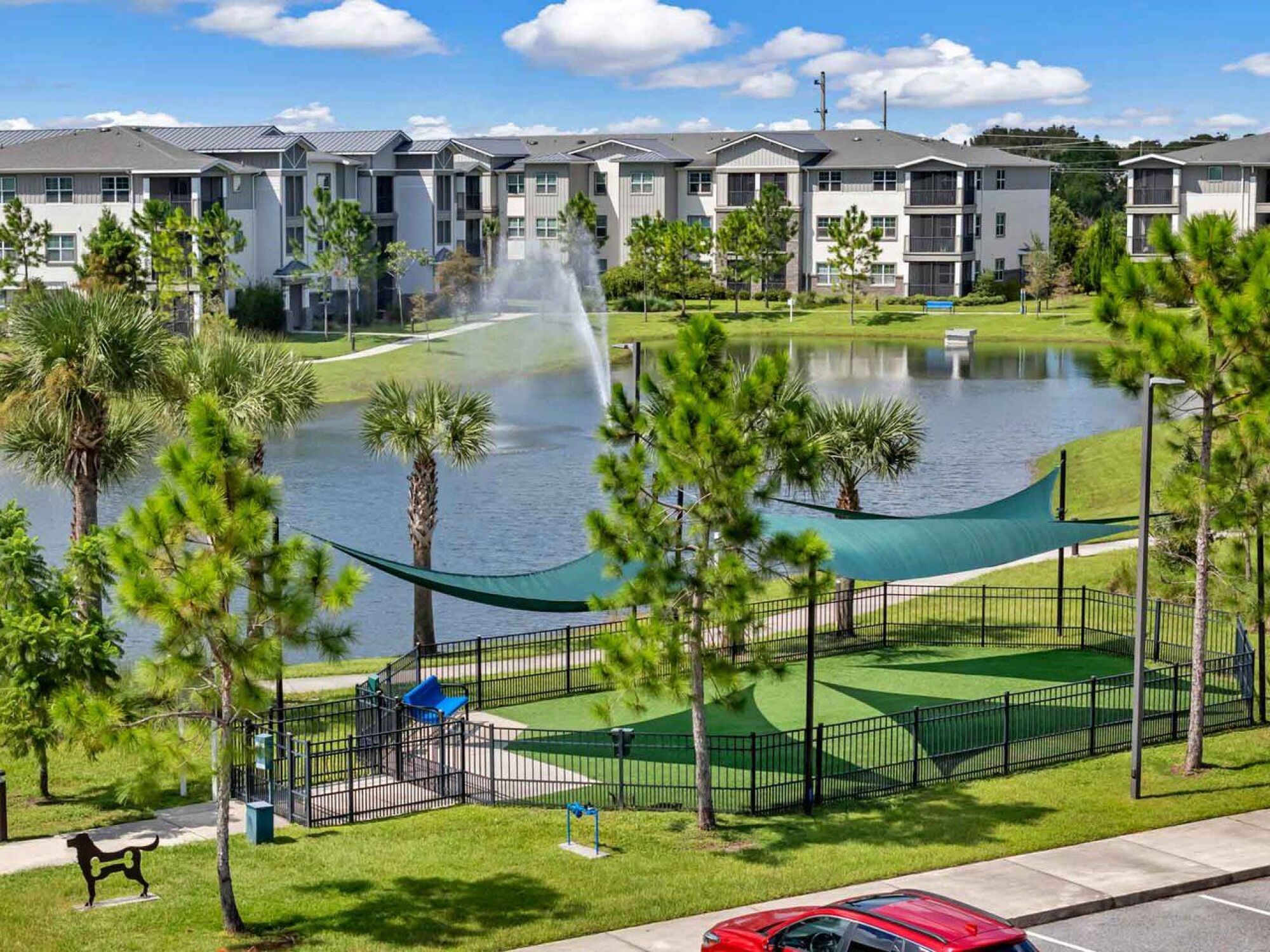 A fenced dog park with shade sails by a pond, fountain, and modern apartment buildings in the background at Luminary at 95 apartments.