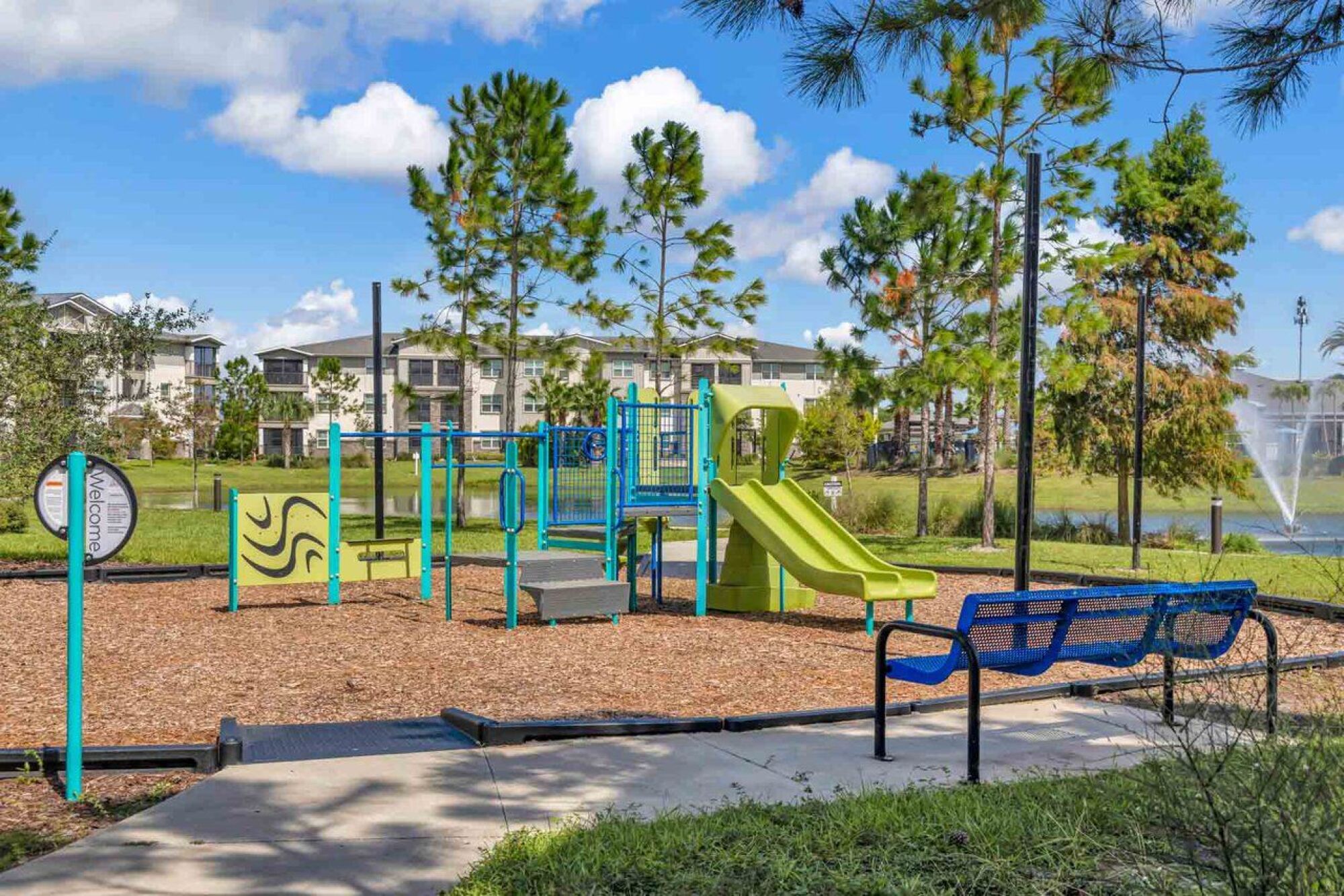 Small playground with slides and climbing bars, surrounded by trees and benches, apartments in the background at Luminary at 95 apartments.