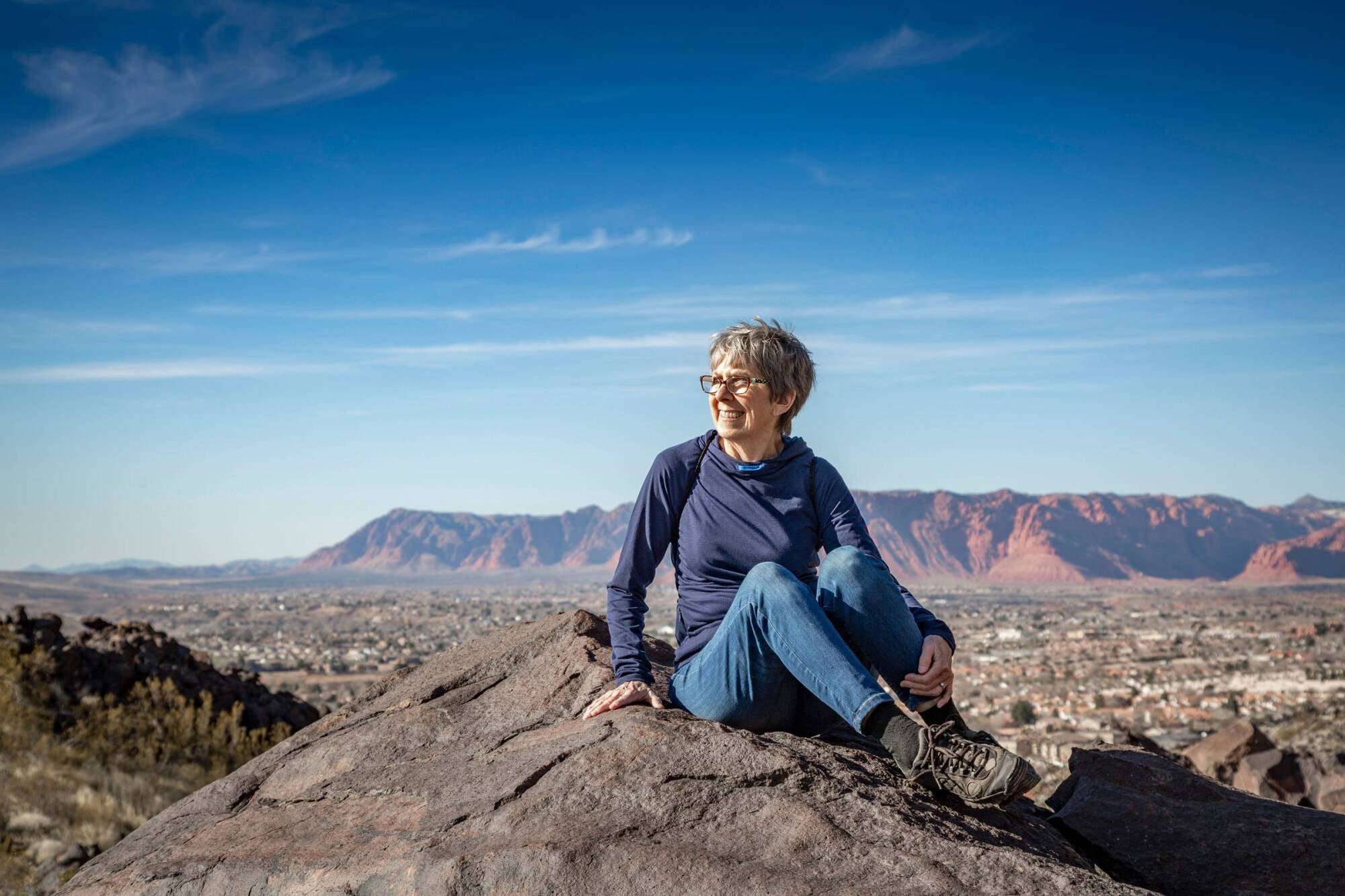 Smiling woman with gray hair sits on a rock, overlooking a city and red mountains under a blue sky.