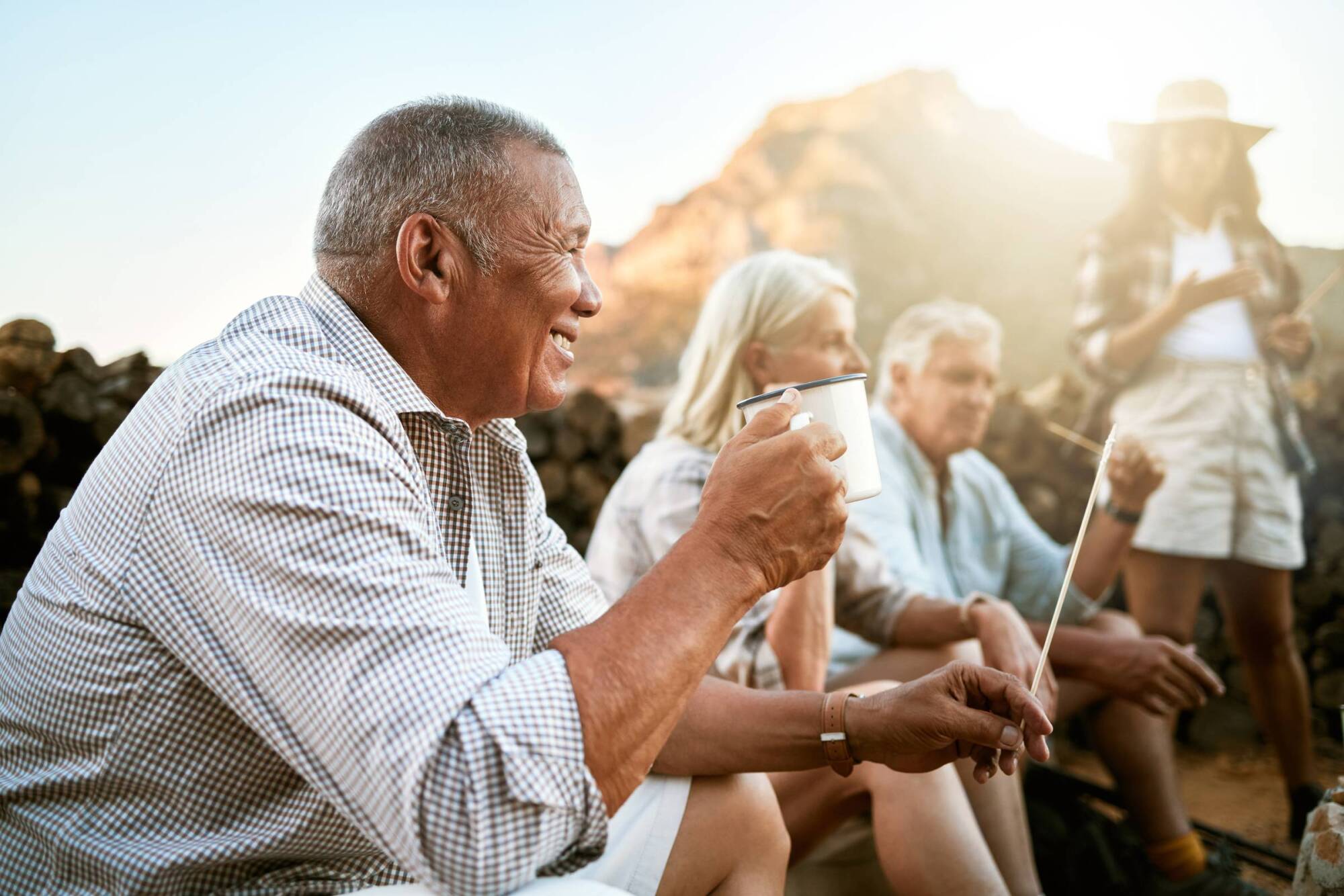 Older adults smile and enjoy drinks outdoors, sitting together near a campfire with mountains in the background.