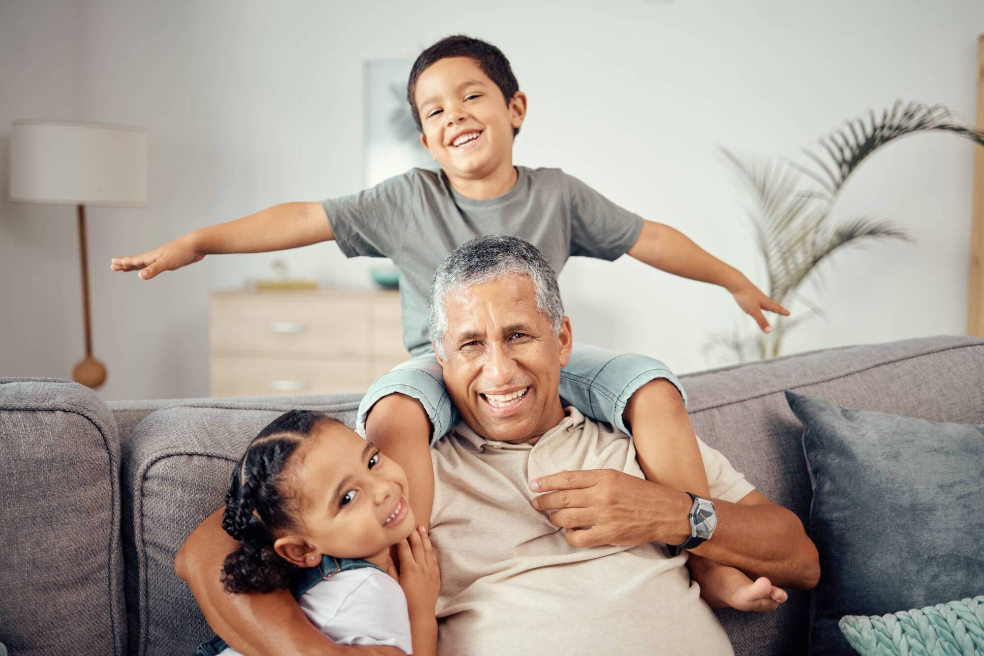 Smiling grandfather sitting on a couch with his two happy grandchildren, a boy and a girl, hugging him.