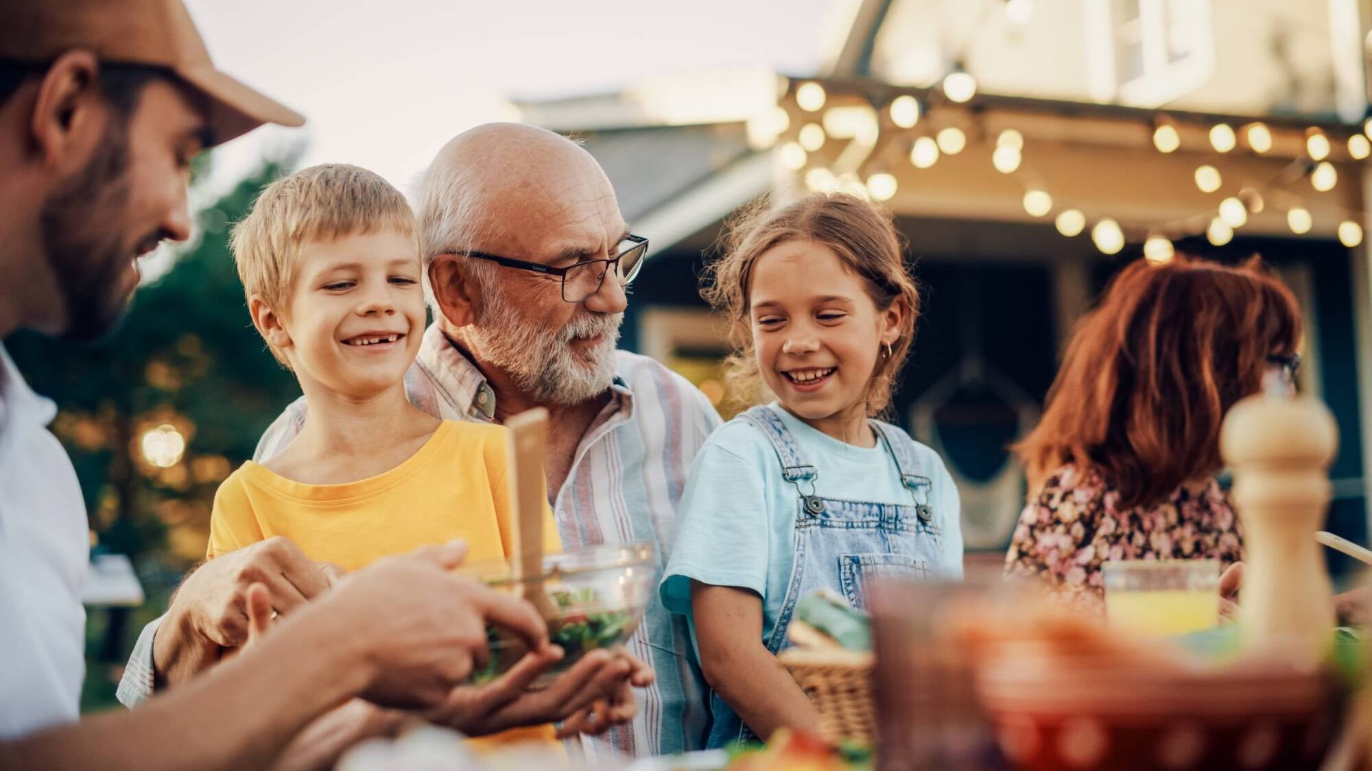 Smiling family enjoys an outdoor meal together in a backyard with string lights hanging above.