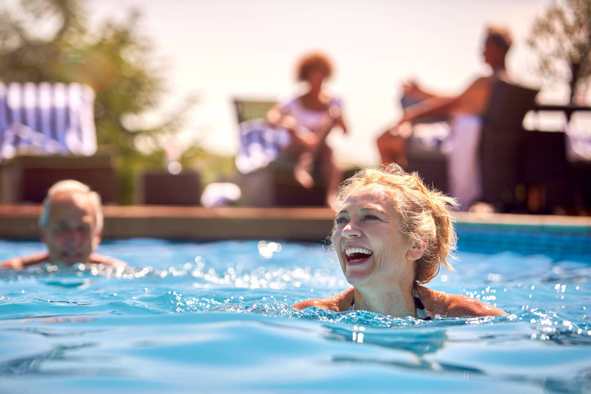 Smiling older woman swimming in a pool, with other people relaxing by the poolside in the background.