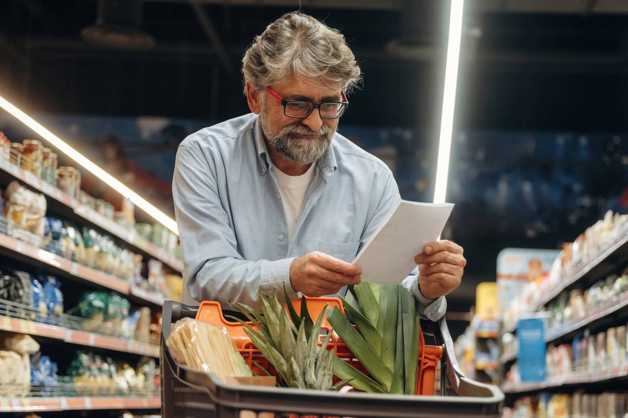 A man with glasses checks a shopping list while pushing a grocery cart in a supermarket aisle.