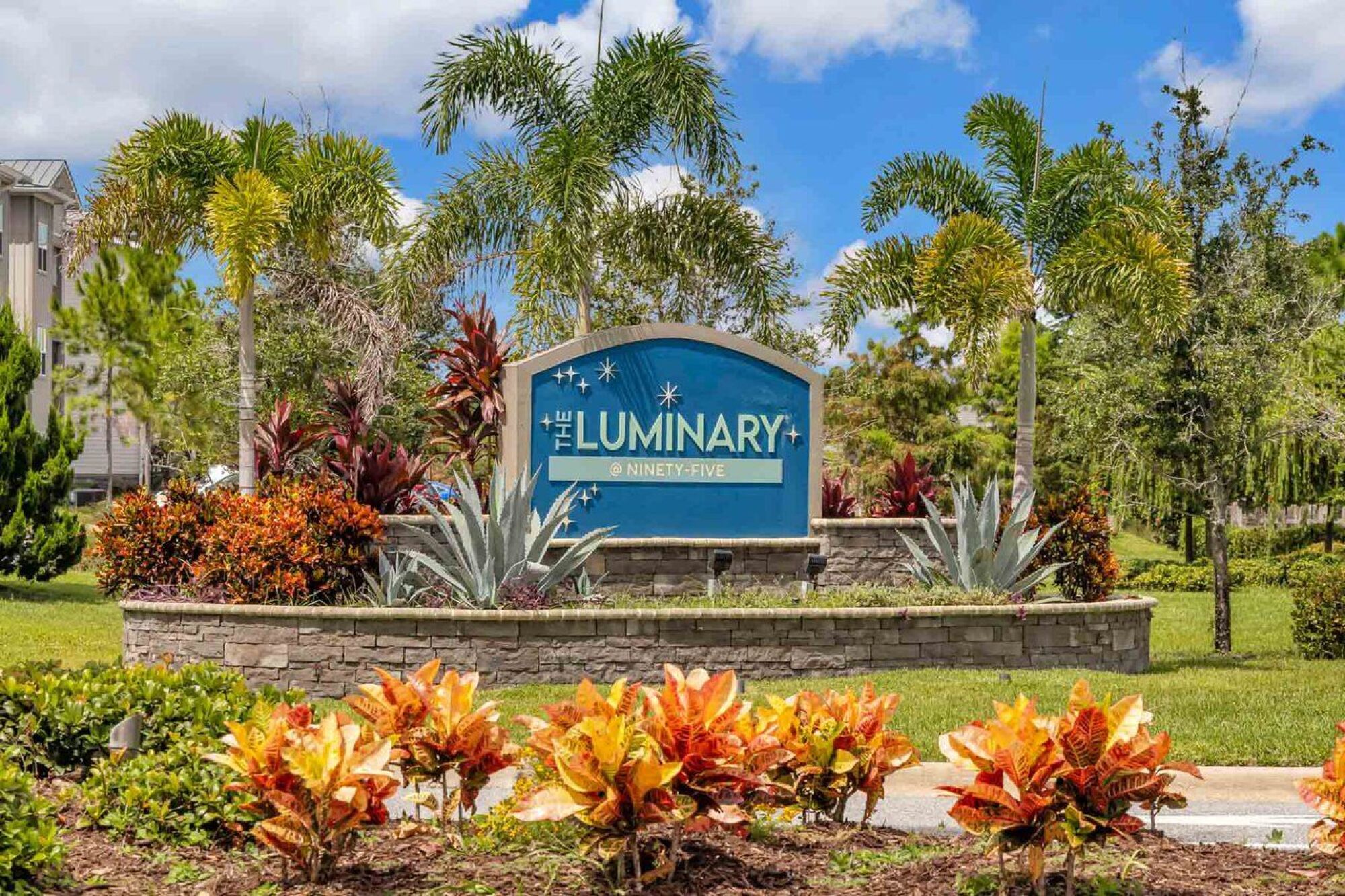 Blue sign reading "The Luminary at Ninety-Five" surrounded by tropical plants and palm trees.