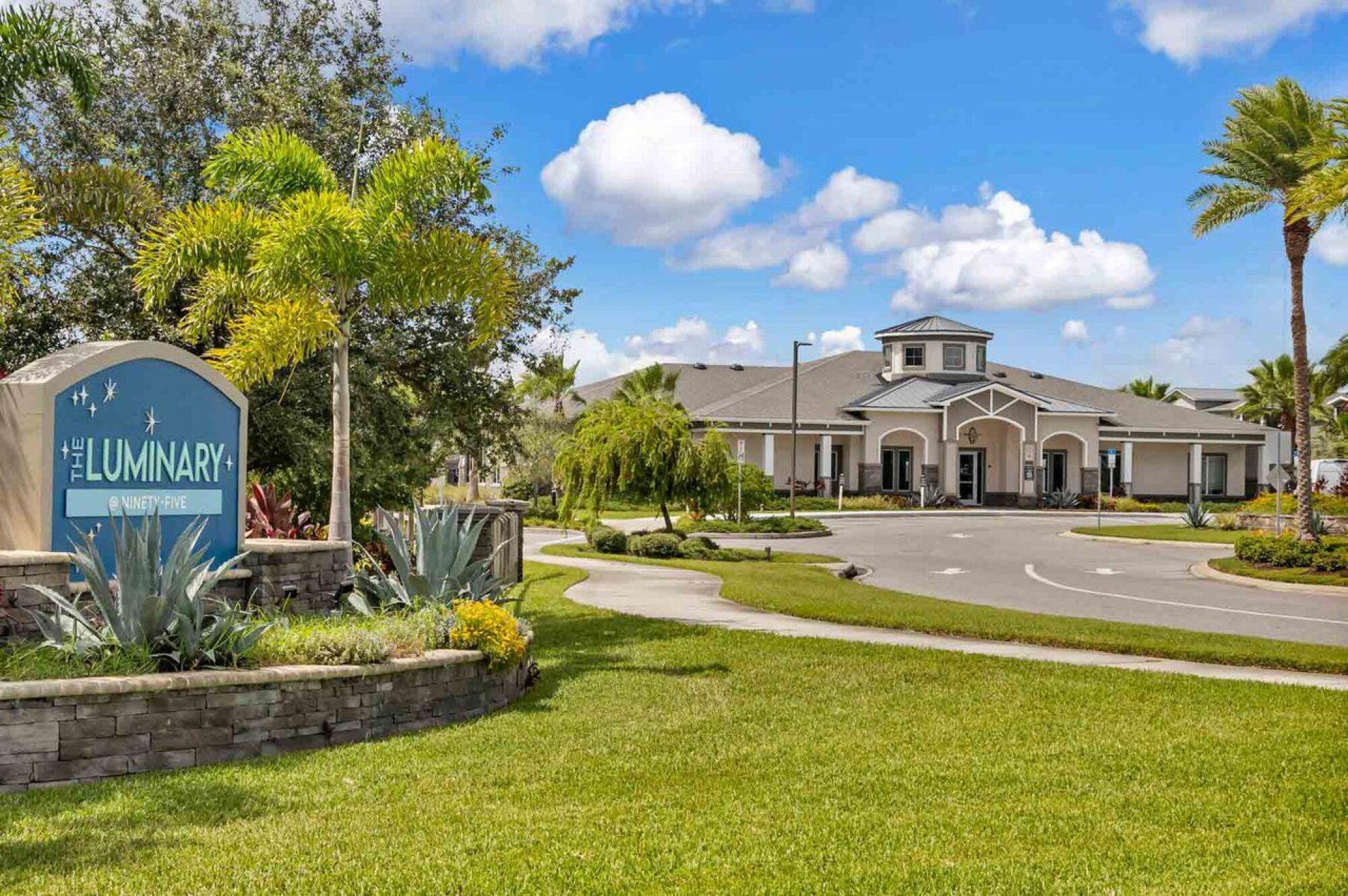 A modern building with arched windows, palm trees, and landscaped grassy areas under a blue sky with clouds at Luminary at 95.